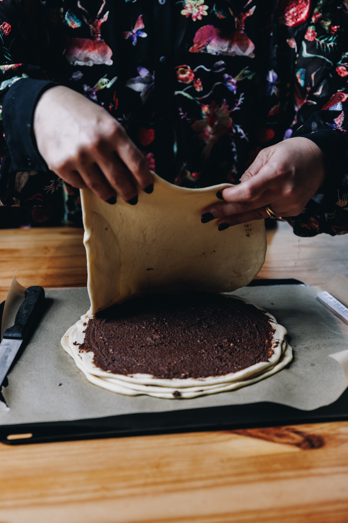 On a black tray with brown baking paper sits a large circle of dough, it is having chocolate spread spread on to it. It is on a wooden table. This is the fourth layer of the Chocolate Star Bread.
