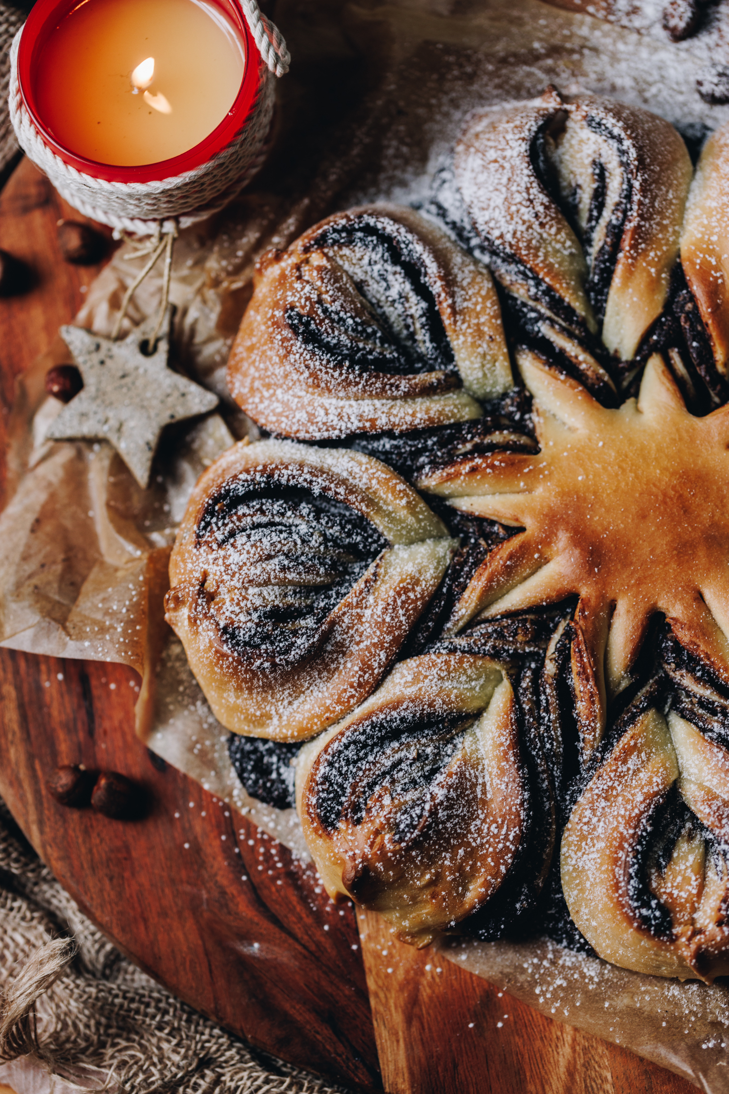 A flat lay of half of a freshly baked chocolate star bread is on a wooden board, dusted with icing sugar and a red candle next to it. It is on a wooden table.
