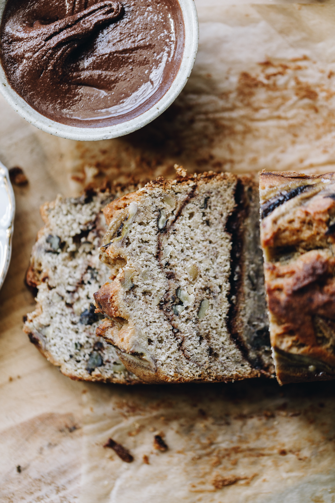 A flat lay shot shows a freshly baked spiced banana loaf that has been sliced and then pieces are laying down on a wooden board. Next to the slices is a small white bowl filled with chocolate almond butter. 
