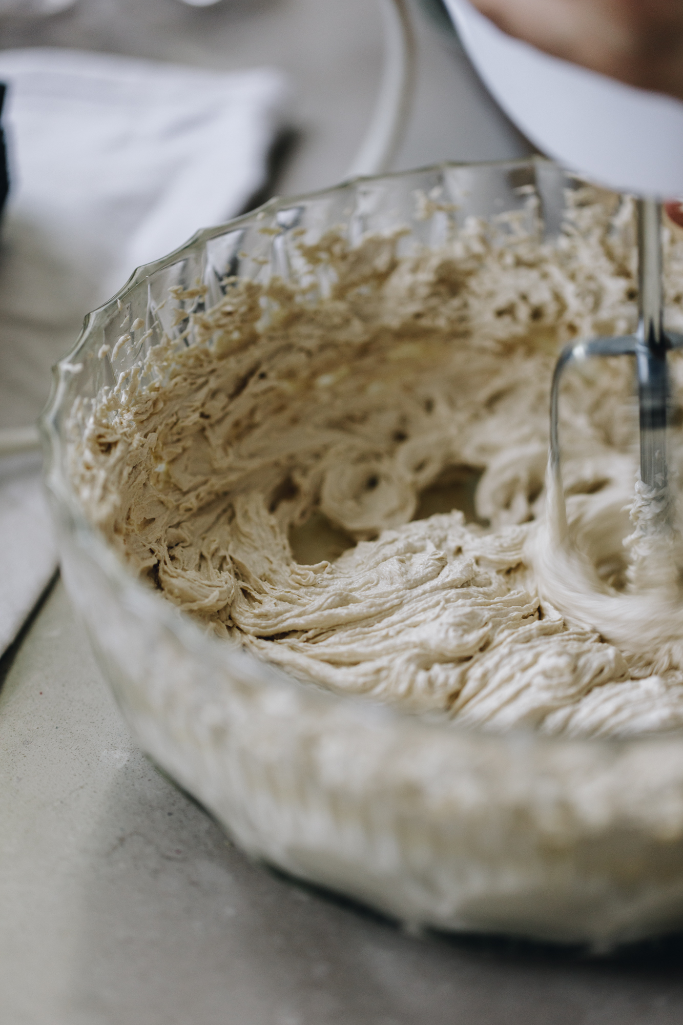 A glass vintage bowl sits on a grey stone bench. In the bowl is whipped butter and sugar that is light in colour and texture. It is being whipped with a white hand mixer. A white cloth is in the background. 