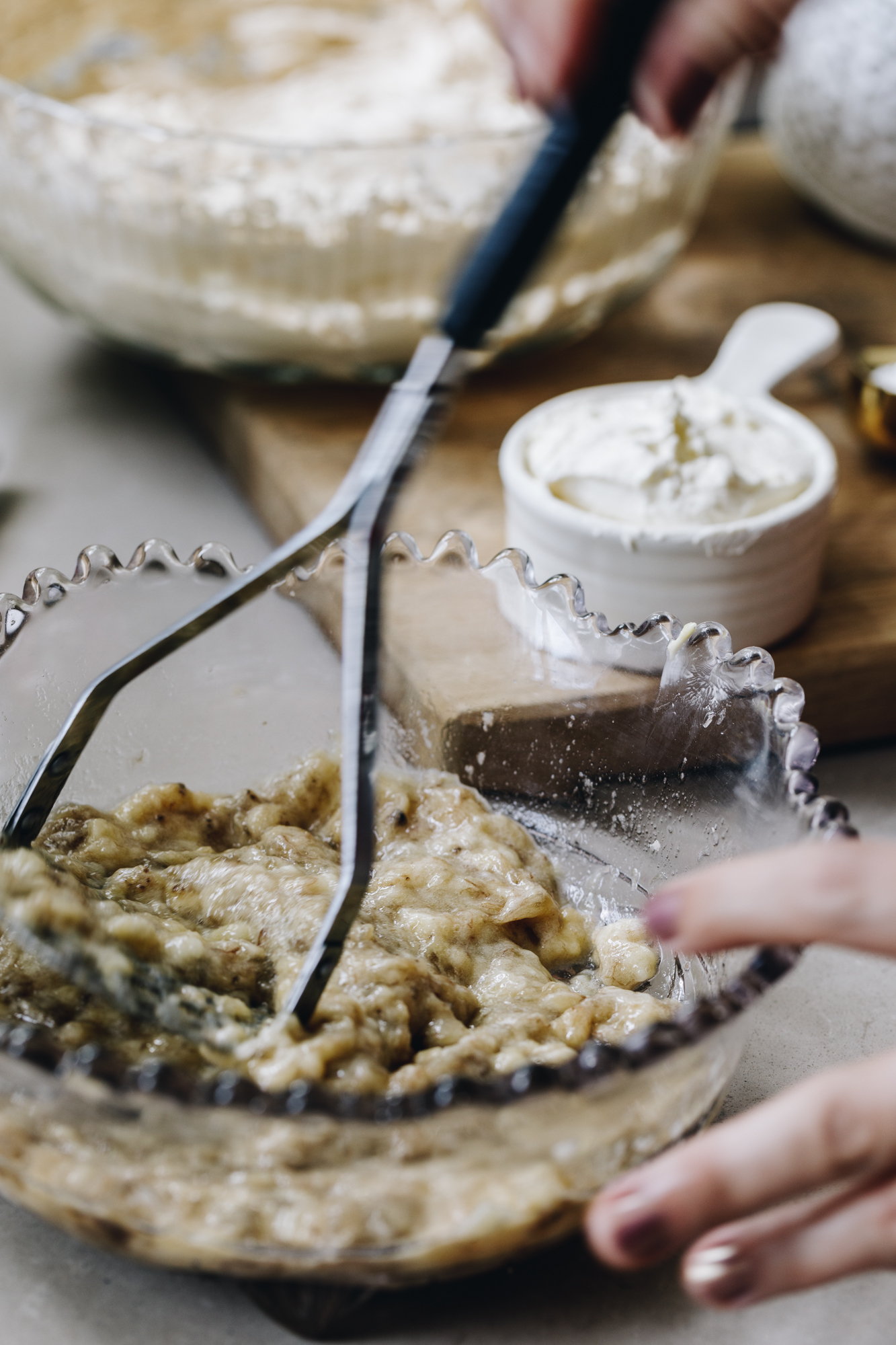 A purple vintage bowl sits on a grey stone bench. In the bowl is mashed bananas that are being mashed with a vintage potato masher. In the background is a wooden board with a white ceramic cup with sour cream in it. A white glass bowl is behind it with whipped butter in it.