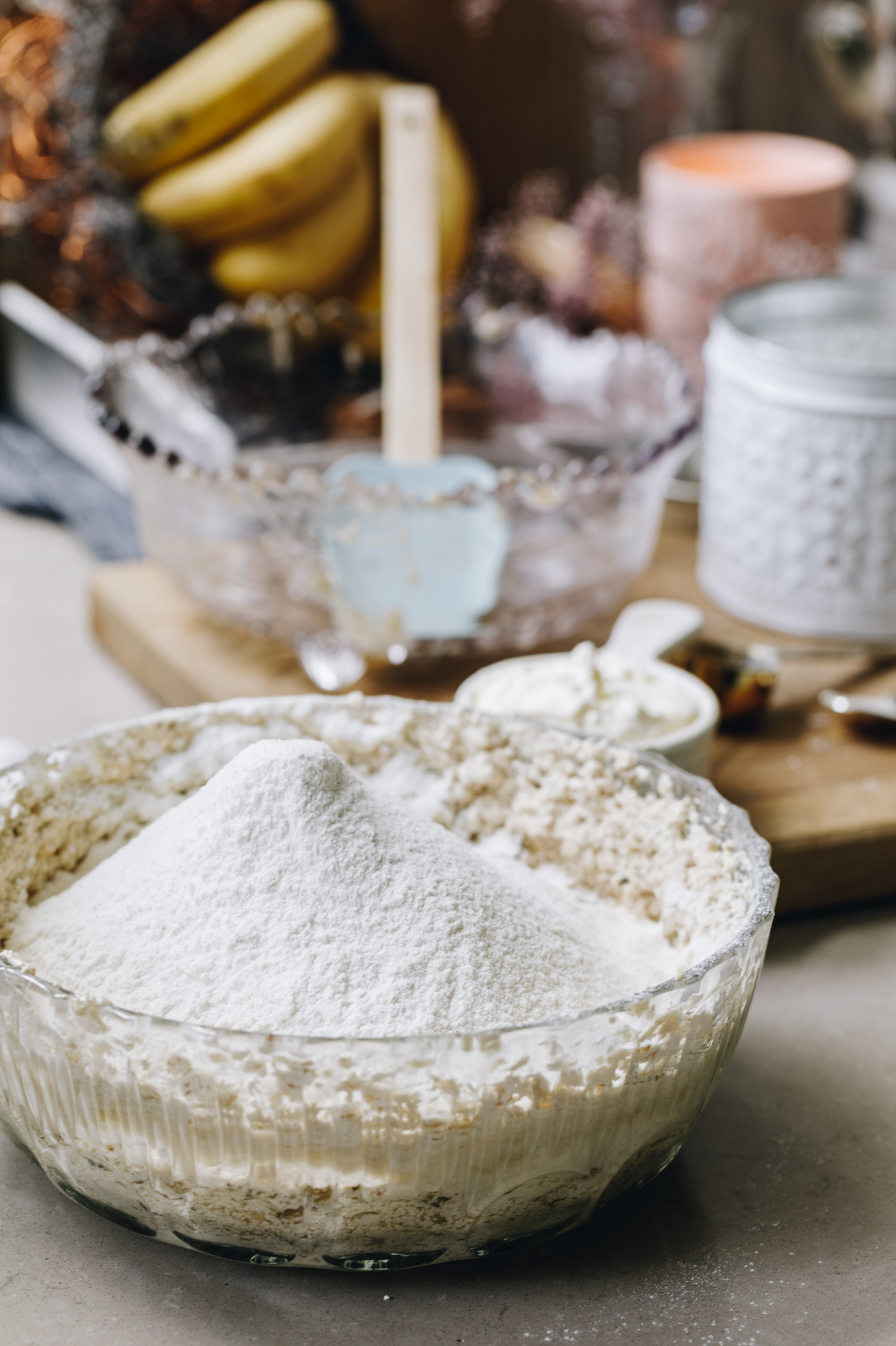 A glass vintage bowl sits on a grey stone bench. In the bowl is sifted dry ingredients that are a mountain shape. Behind the bowl is another vintage bowl on a wooden board with a spatula in it. A white metal vase is next to it and a bunch of yellow bananas and a peach candle are in the background.