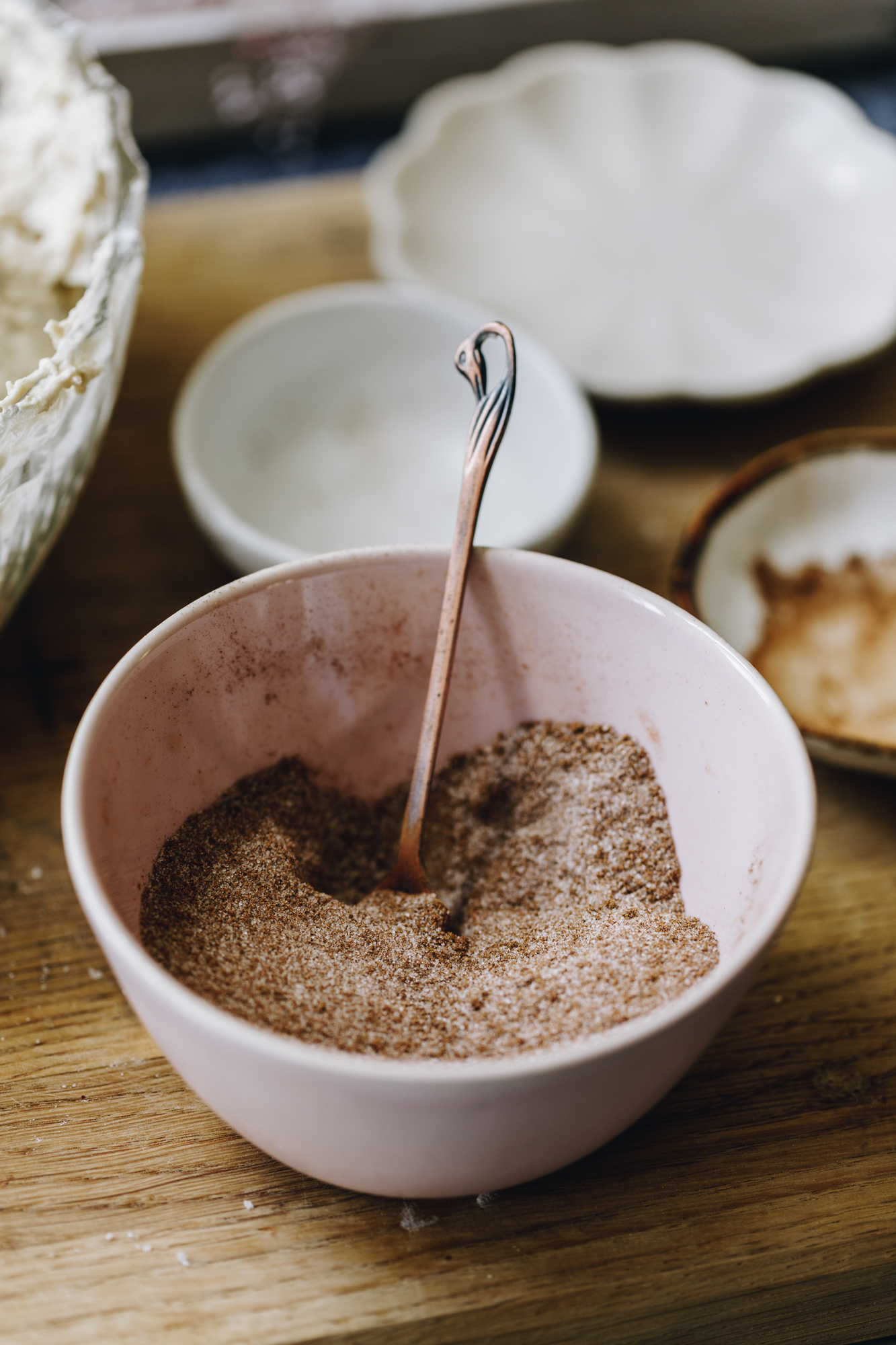 A small pink bowl sits on a wooden board. In the bowl is a spiced sugar. In behind it on the board are white small bowls. 