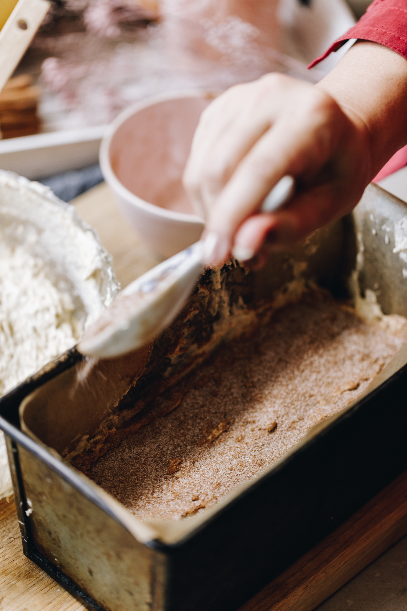 A vintage loaf tin lined with brown baking paper sits on a wooden board. In the tin is banana loaf mixture. Naomi is using a ceramic spoon to sprinkle over the spiced sugar mixture on top. A glass bowl and a white bowl are in the background. 