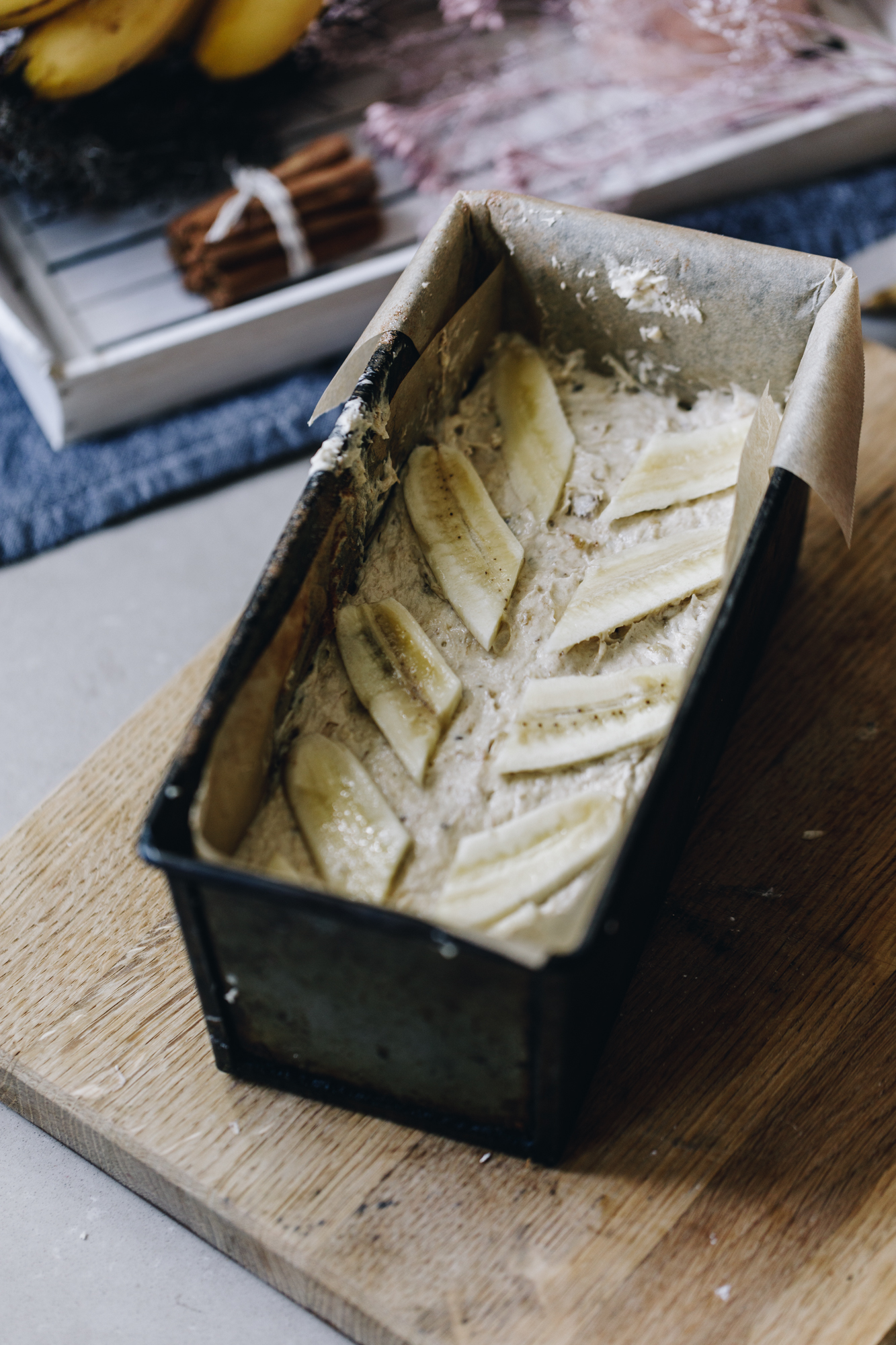 An unbaked spiced banana loaf is in the tin and decorated with slices of banana. It sits on a wooden board. In the background is a white tray on a blue cloth. On the white tray is a small stack of cinnamon sticks that are tied in string along with a bunch of bananas.