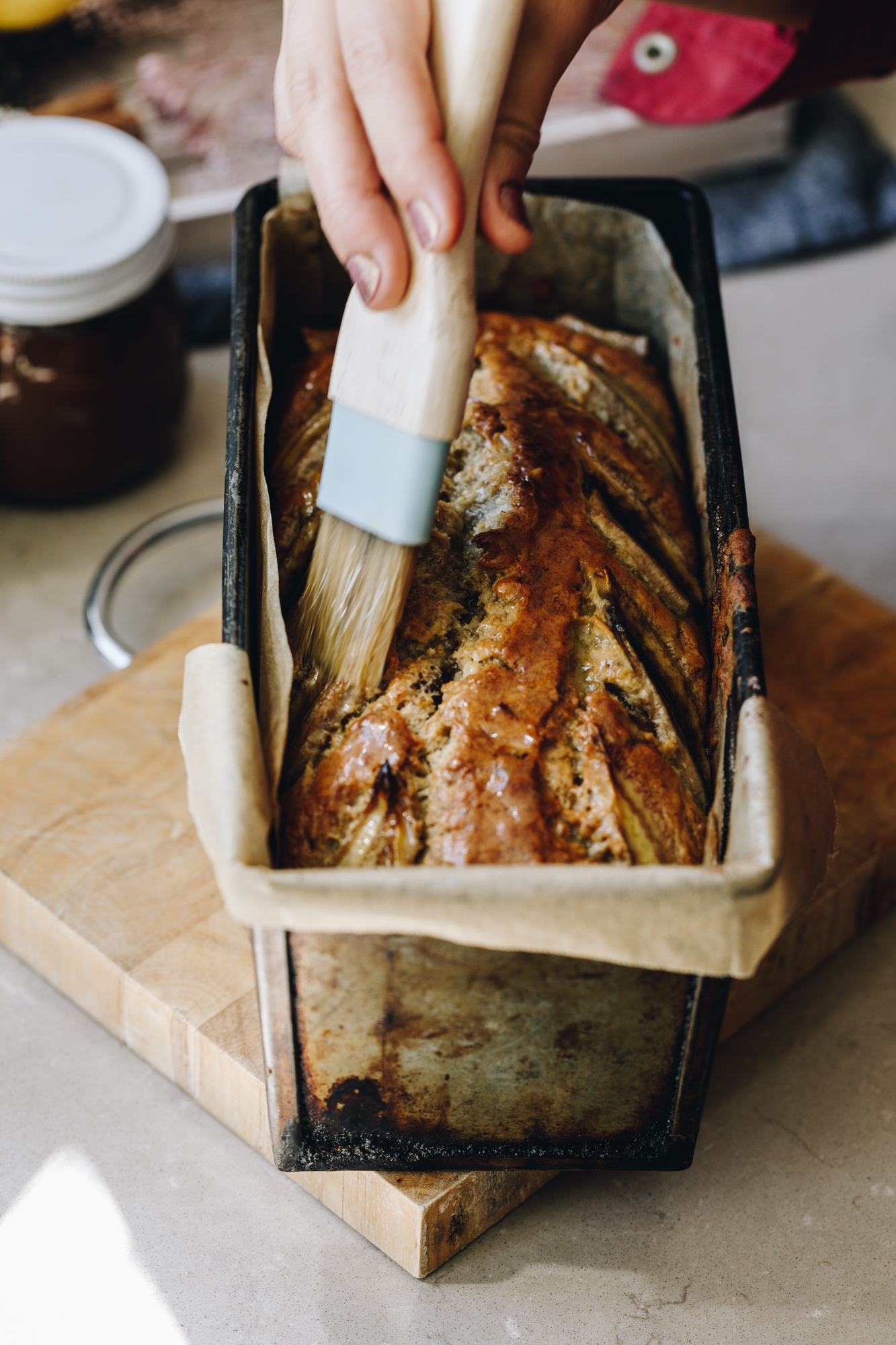 A freshly baked spiced banana loaf is on a small wooden board, on a grey stone bench. The loaf is being glazed with butter by Naomi with a wooden pastry brush. A small jar of chocolate almond butter is in the background. 