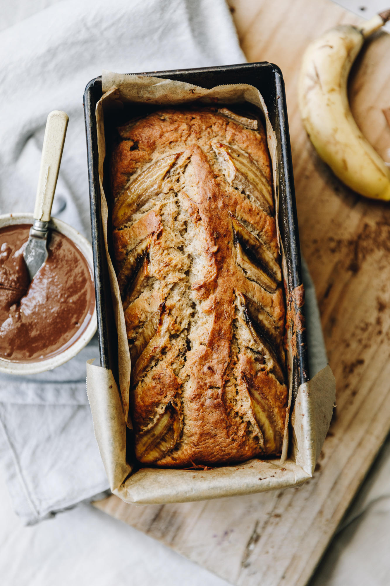 A flat lay shot shows the baked spiced banana loaf  in the tin lined with brown baking paper. There is a white bowl of chocolate almond butter next to it. The tin sits on a blue tea towel. A whole banana is next to it. 