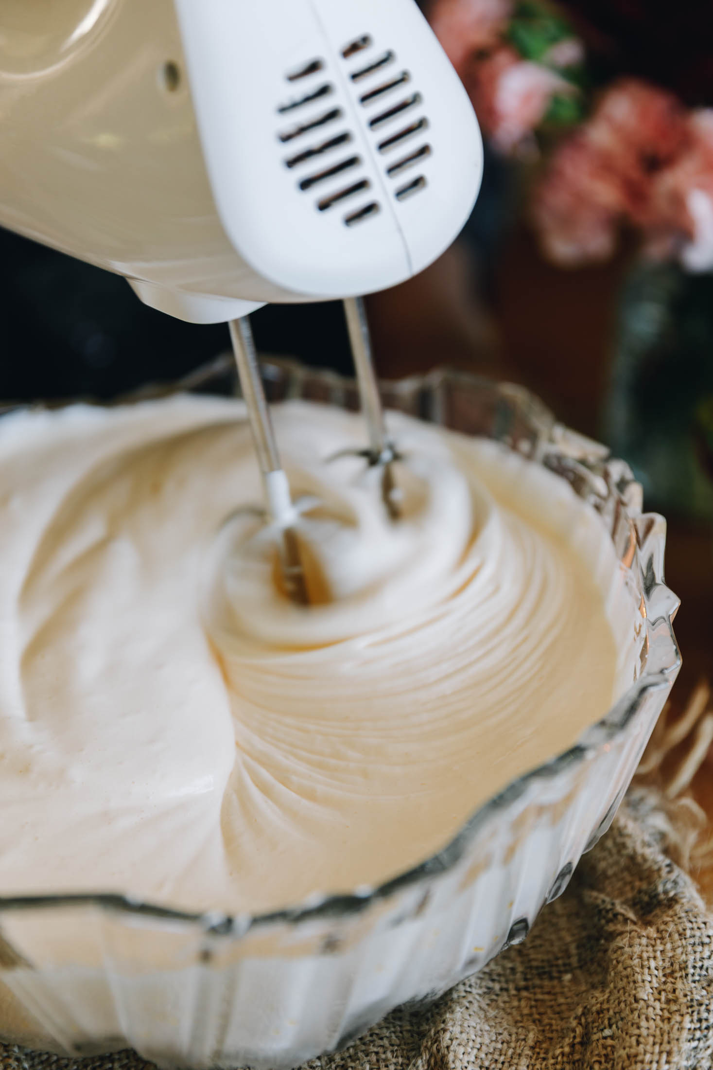 A glass vintage bowls sits on a piece of natural fabric. In the bowl is whipped eggs and sugar that are being whipped with a white hand mixer.