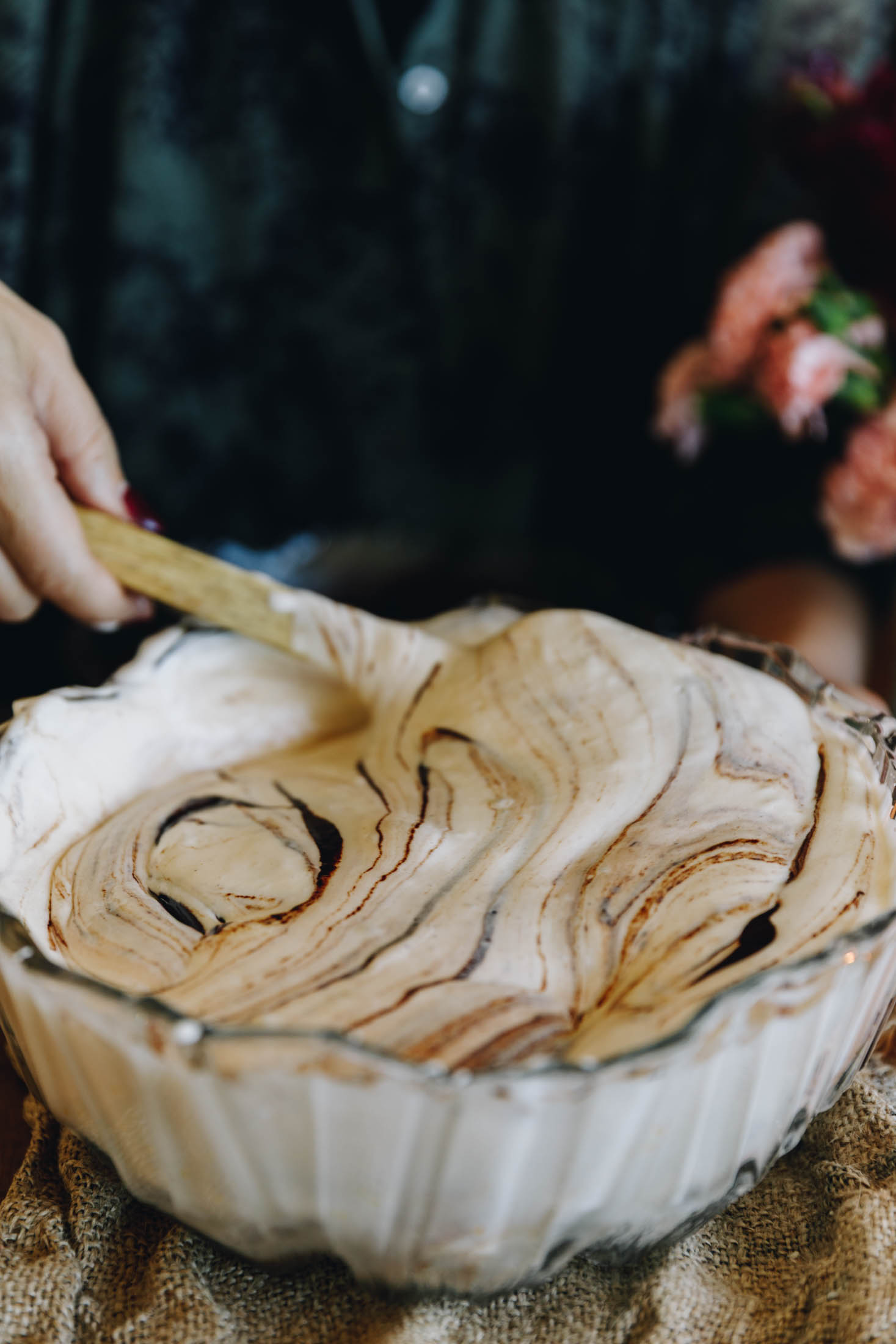 A glass vintage bowls sits on a piece of natural fabric. In the bowl is whipped eggs that have chocolate ganache swirled through it. Naomi is using a spatula to fold it in.