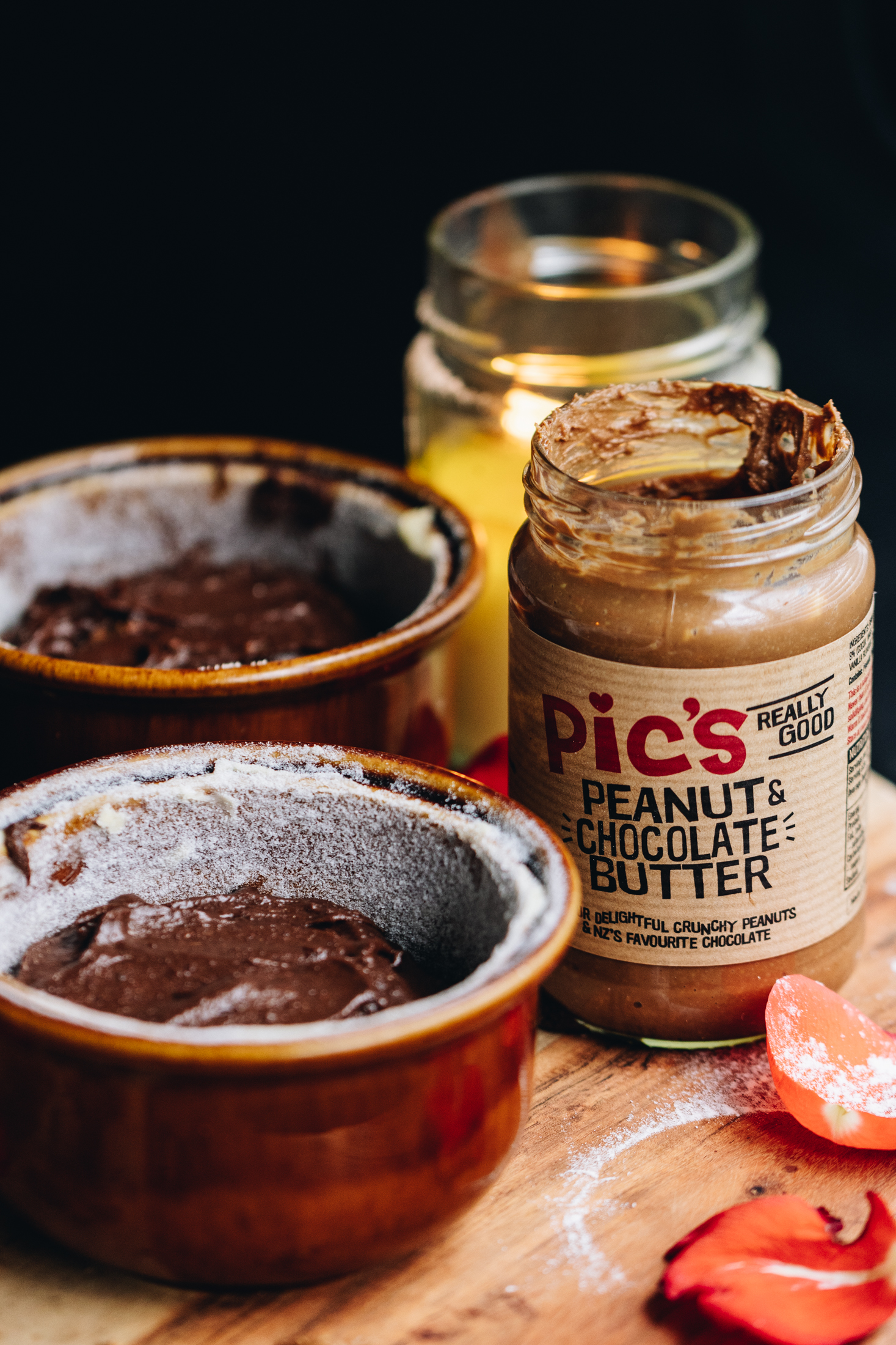 On a wooden board sites too small brown ramekins that have the lava cake batter in it, they are not baked. Next to the ramekins is a jar of PIc's chocolate peanut butter and behind it is a yellow candle burning in a jar. Two rose petals are on the board too.