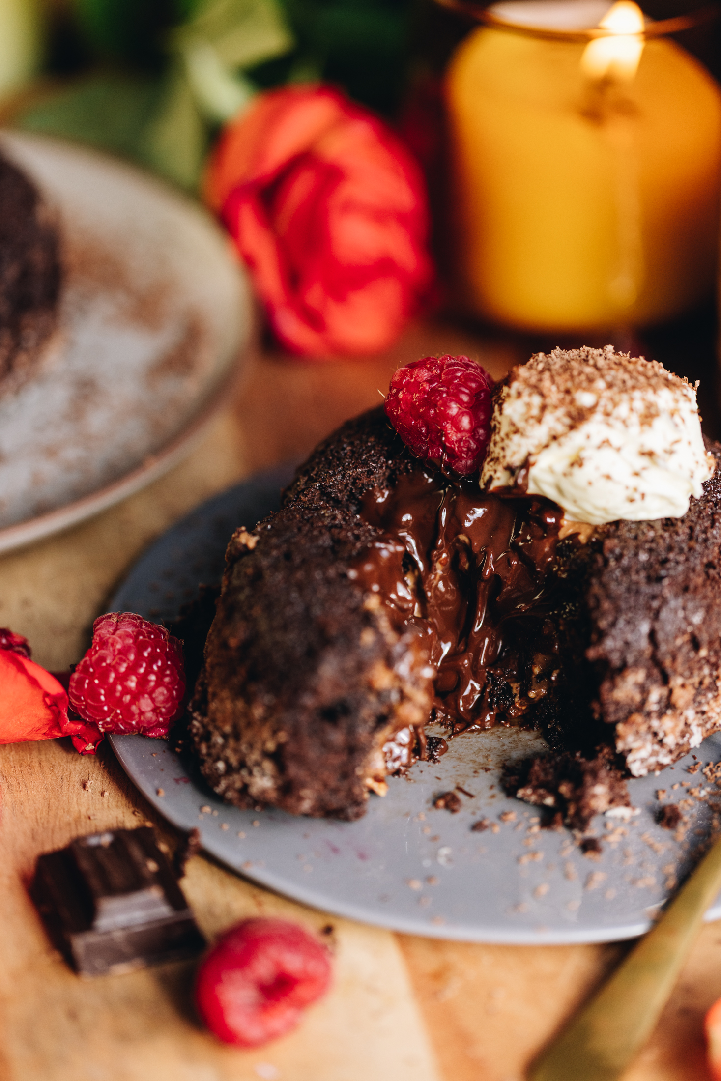 A close up shows a baked chocolate pudding (lave cake) that is on a small grey plate. The lava cake has had the front of it removed to show to gooey chocolate centre. In the background a yellow candle is burning with an orange rose next to it. Fresh raspberries, a piece of dark chocolate and orange rose petals are in front of the grey plate. Another plate can be seen on the side of frame.