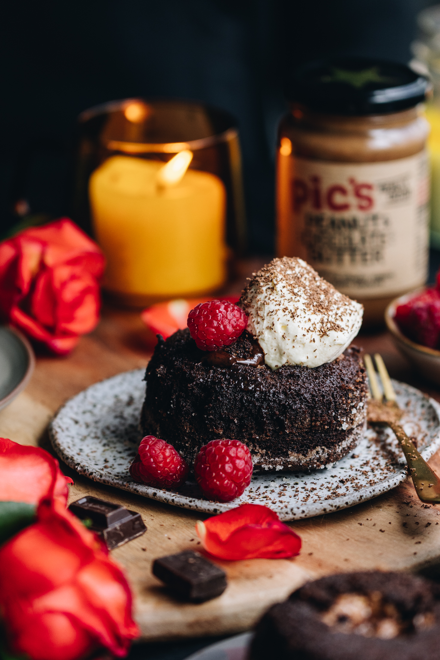 A baked gooey chocolate lava cake is on a grey ceramic plate with a small fork next to it. It is whole and has whipped cream on top of it with a fresh raspberry and grated chocolate. There are two raspberries on the plate next to the cake. Around the edge of frame is orange rose petals. Behind the cake is a yellow candle burning in a glass jar with an orange rose next to it. On the other side is a blurred pics chocolate peanut butter jar.