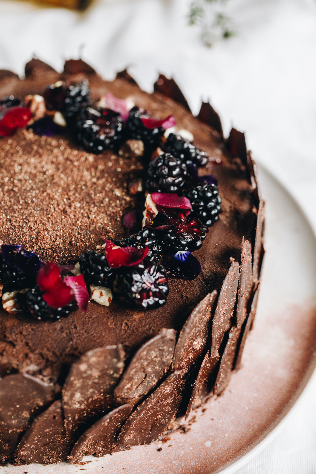 A freshly decorated vegan chocolate cake sits on a pink plate on top of a white table cloth. The chocolate cake is wrapped in dark chocolate decorations and has blackberries and pink flowers arranged on the top.