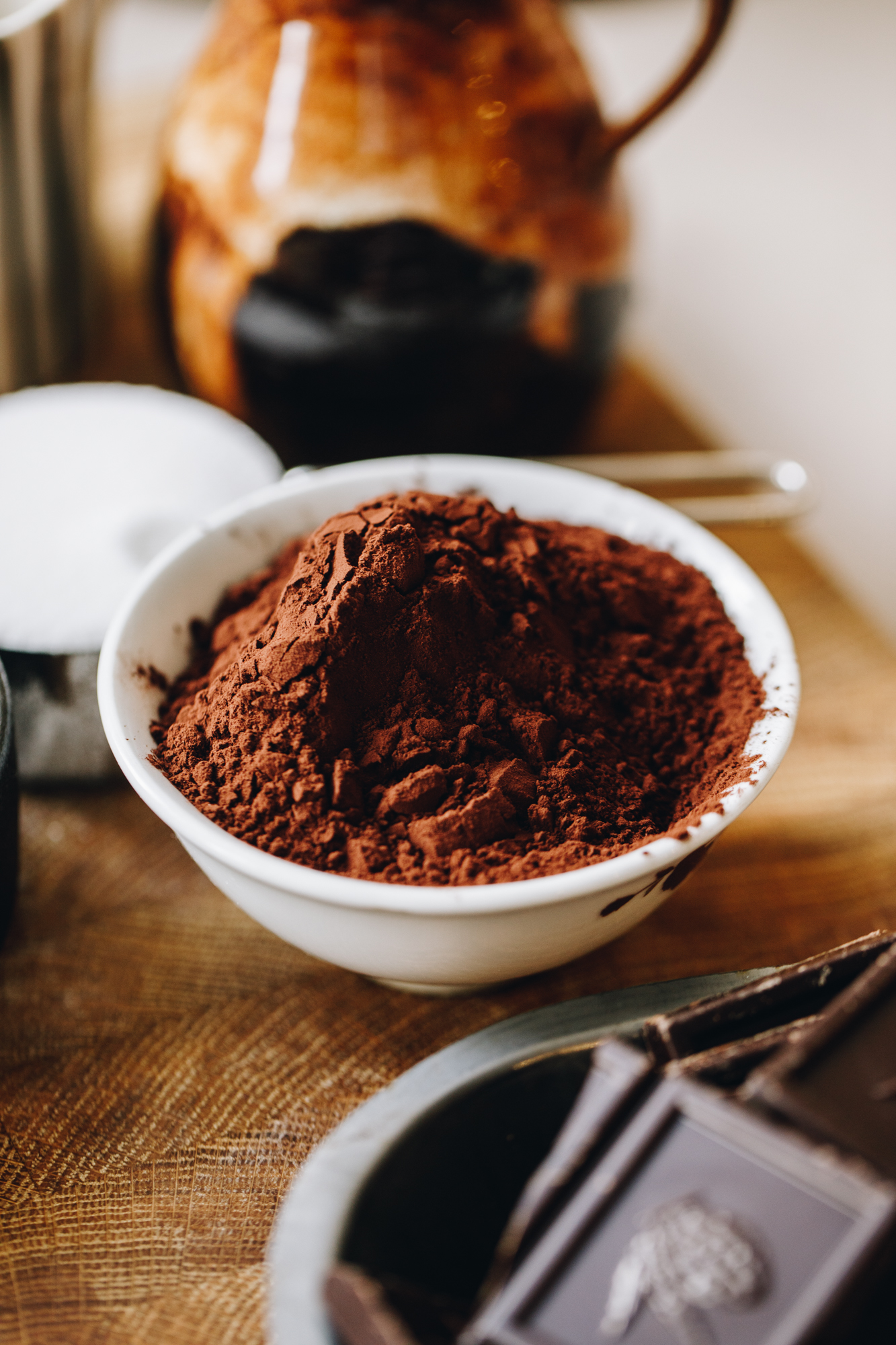 A wooden board has chocolate in a silver container, cocoa in a white bowls, milk in a silver cup and a black and brown jug in the background. 