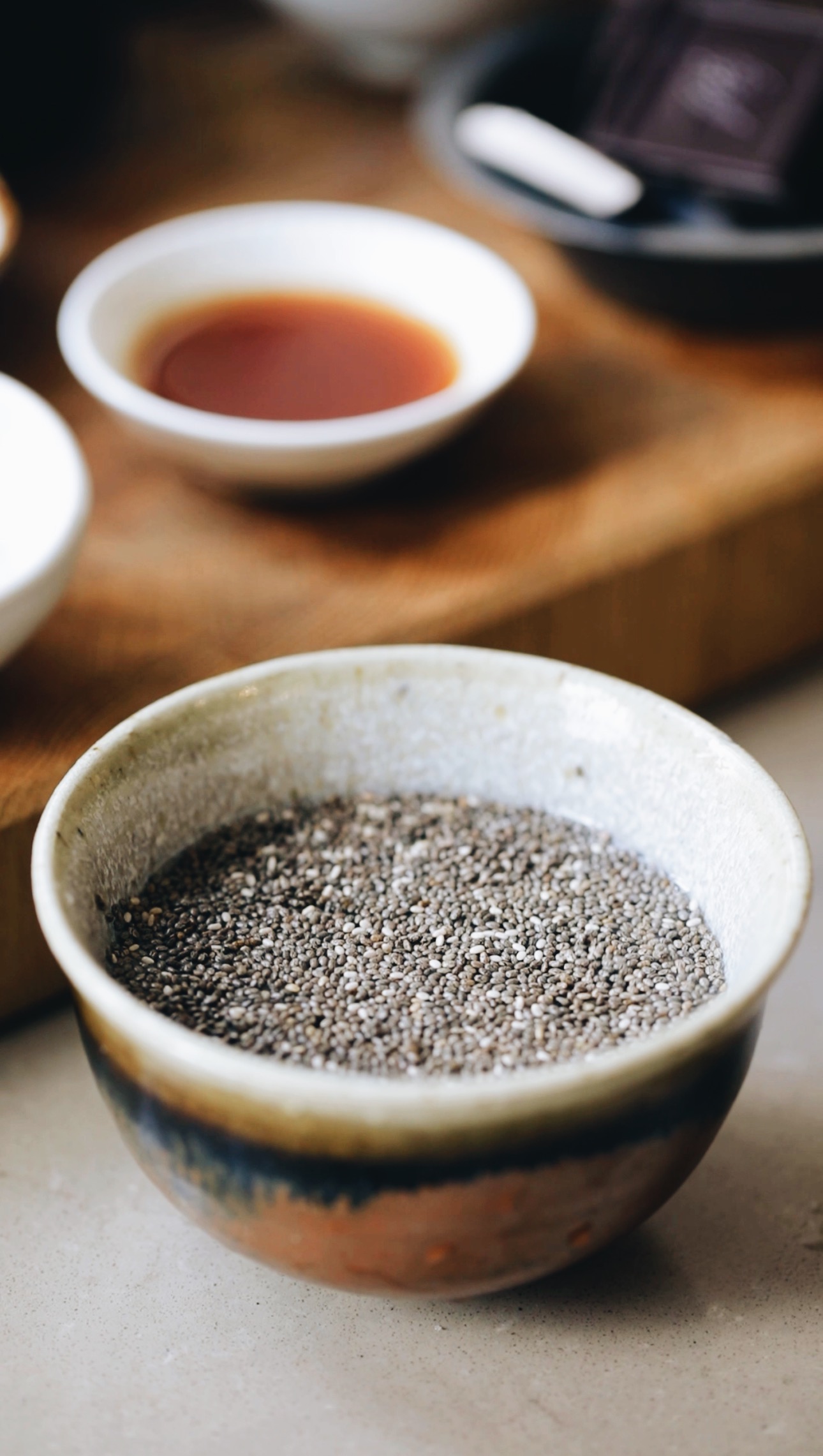 A small brown ball sits on a brown stone bench with chia seeds and water on it. Behind the bowl is a wooden board with a small white bowl with vanilla in it. 