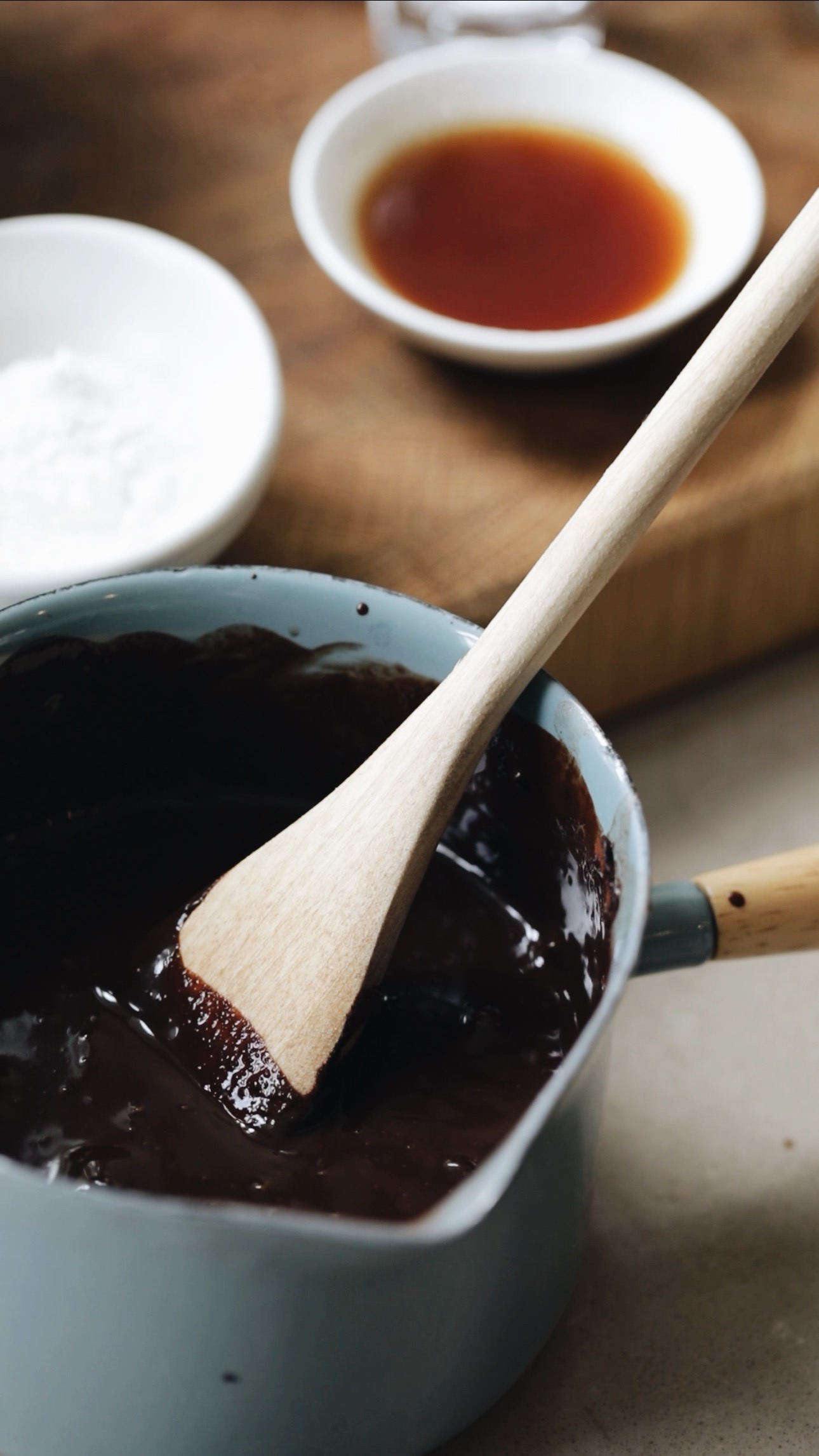 A small green pot has melted chocolate in it. Behind the bowl is a wooden board with a small white bowl with vanilla in it.