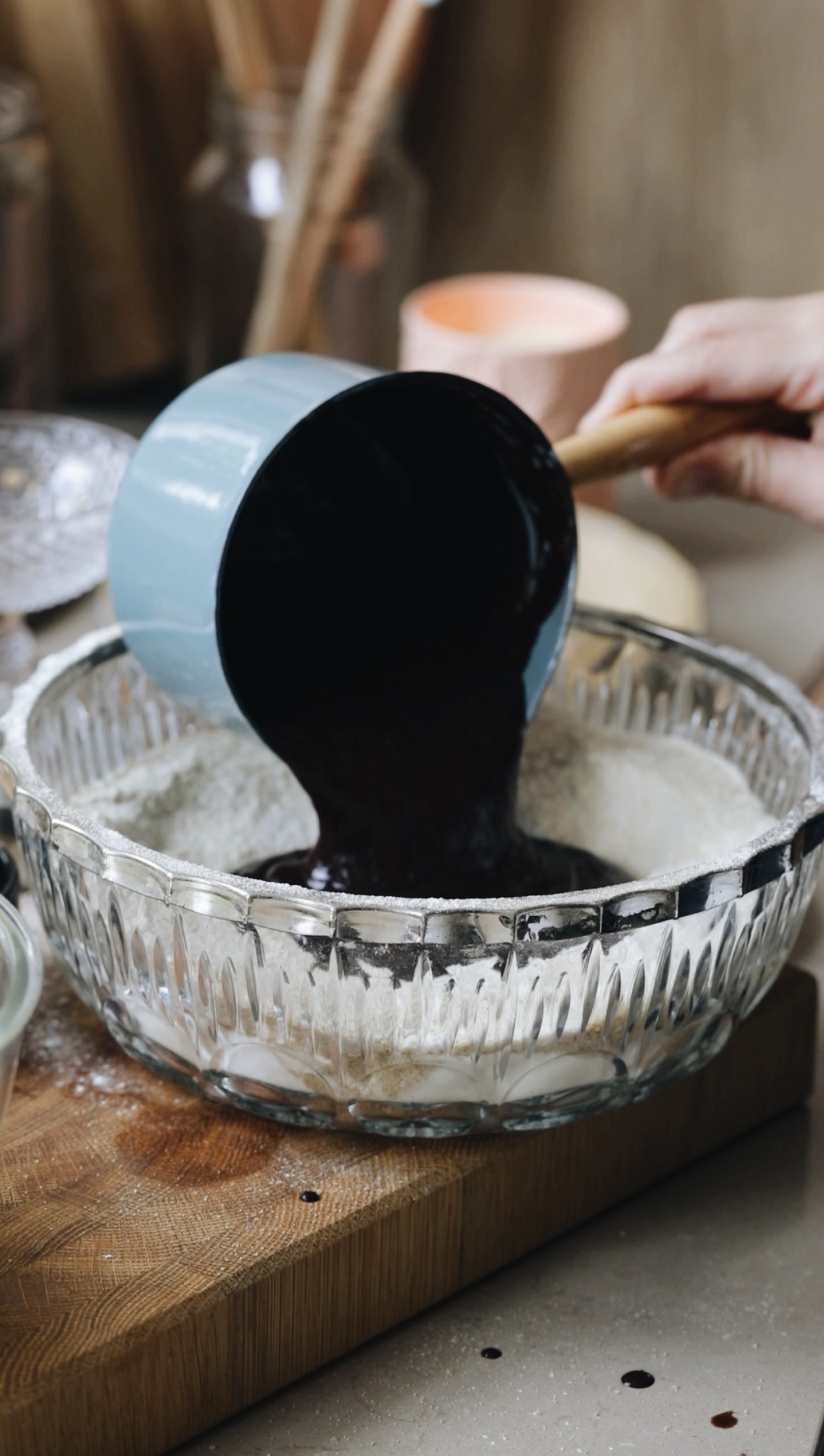 A vintage glass bowl sits on a wooden board. The bowl has dry ingredients in it and Naomi is pouring a chocolate mixture in to it with a green pot. There is a peach candle in the background. 