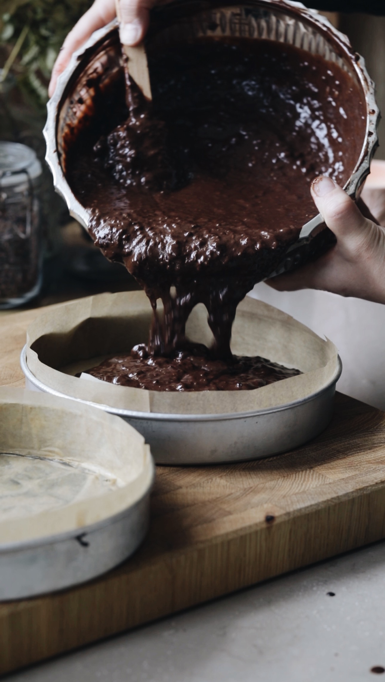 Two silver cake tins lined with brown baking paper sit on a wooden board. Naomi is pouring chocolate cake batter in to one of them from a glass vintage bowl. 