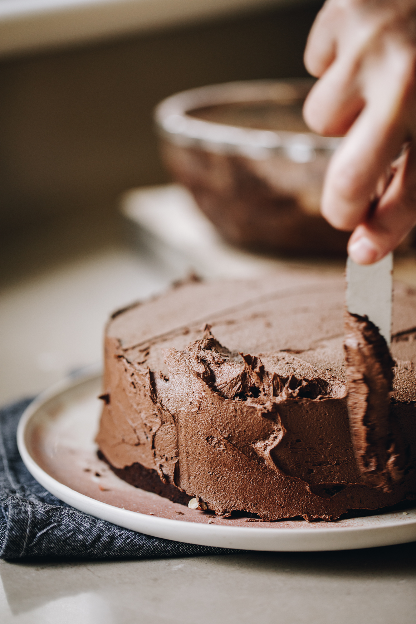 A chocolate vegan cake is on a pink plate, that has a blue tea towel under it, on a brown stone bench. Naomi is smearing the cake with vegan chocolate buttercream using a knife.
