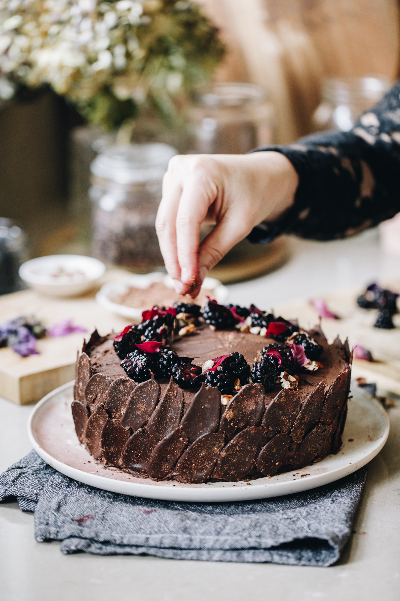 A freshly decorated vegan chocolate cake sits on a pink plate on top of a white table cloth. The chocolate cake is wrapped in dark chocolate decorations and has blackberries and pink flowers arranged on the top. Naomi is sprinkling the cake with rose petals. In the background is a small wooden board that has decorating things on it. Flowers are vases are in the background too. 