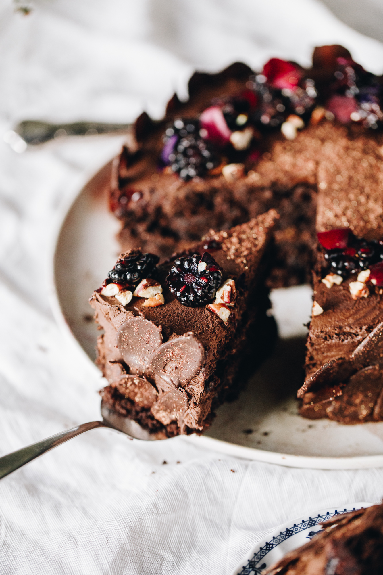 A freshly decorated vegan chocolate cake sits on a pink plate on top of a white table cloth. The chocolate cake is wrapped in dark chocolate decorations and has blackberries and pink flowers arranged on the top. A slice has been cut and sits on a cake server that is on the edge of the plate. 