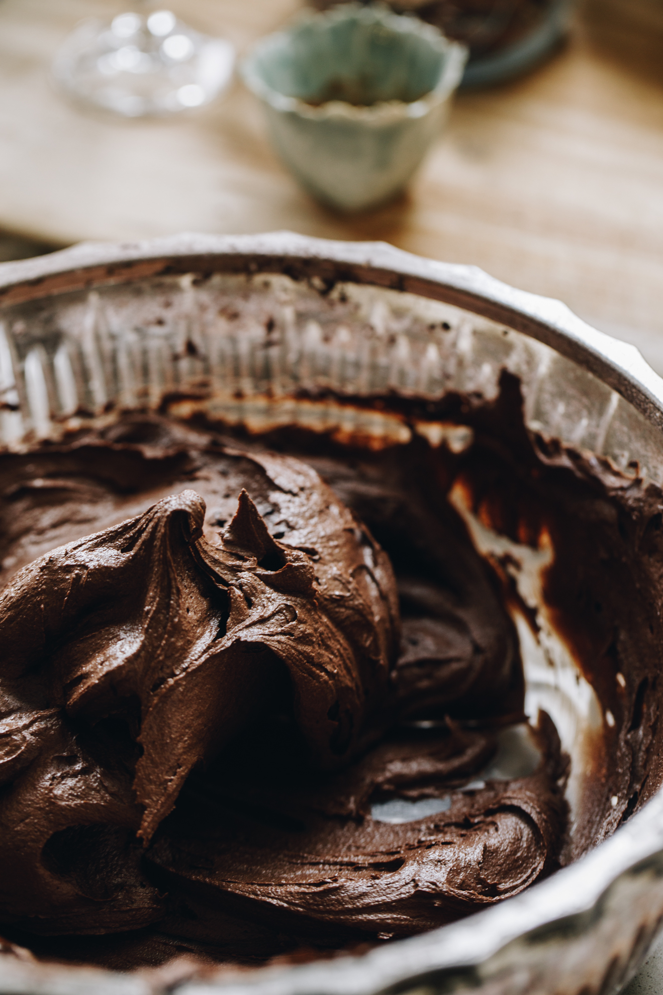 A close up shots shows the vegan chocolate buttercream whipped to perfection in a glass vintage bowl with silver trim. Behind it is a wooden board with small empty vessels on it. 