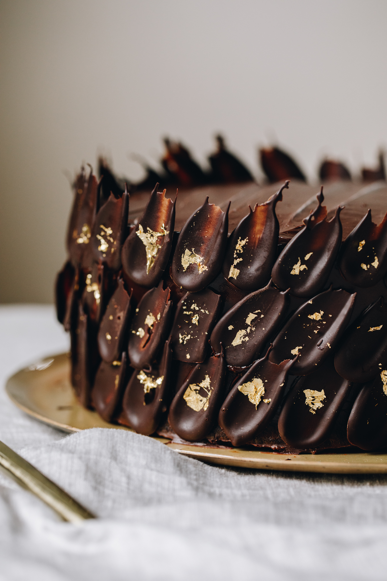 A chocolate cake sits on a golden plate on top of a grey tablecloth. On the sides of the cake are dark chocolate petal decorations that are touched with gold leaf on each one. 
