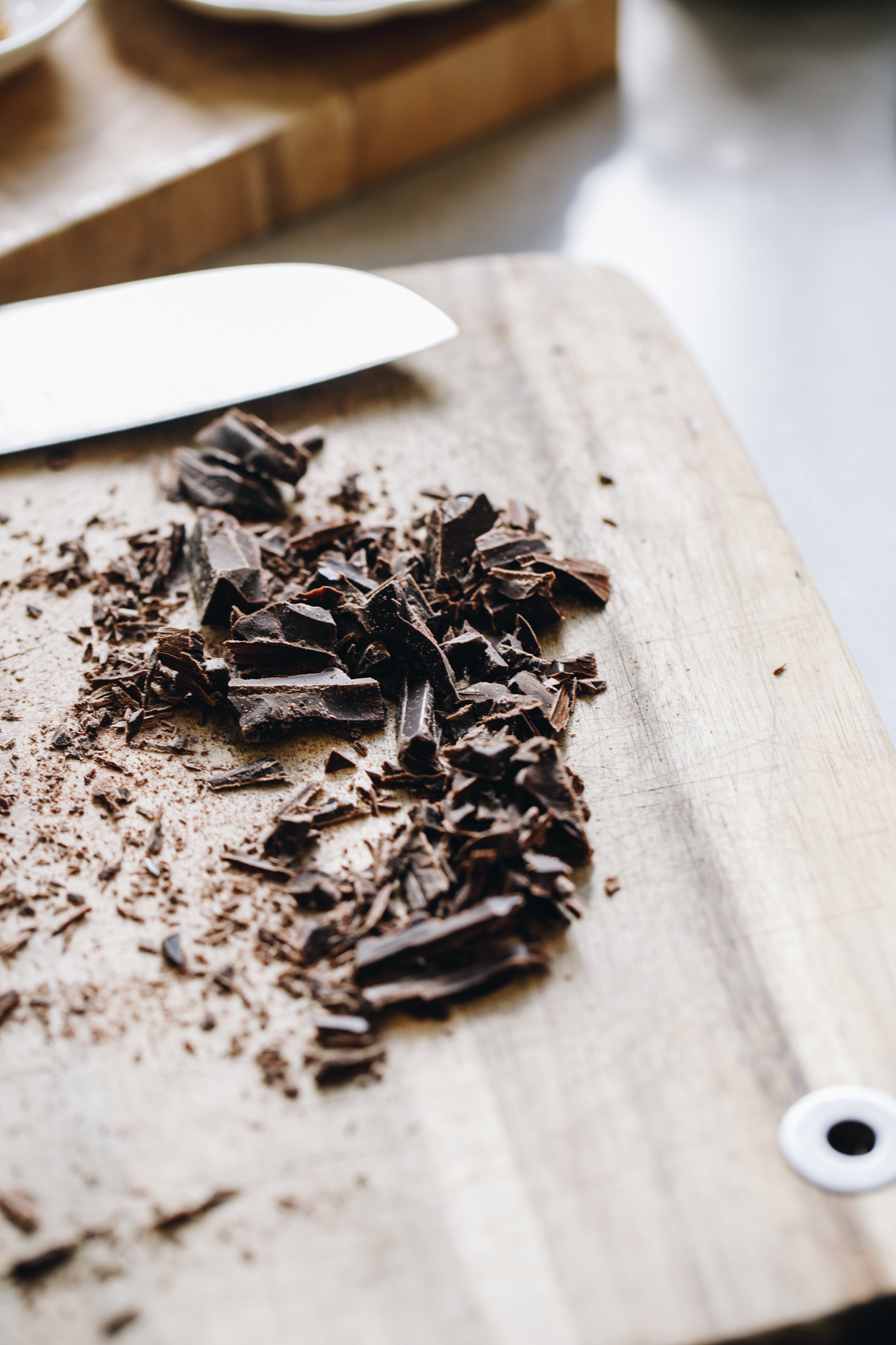 A wooden board sits on a brown stone bench. On the board is the tip of a sharp knife and dark chocolate that has been cut in to shards. 