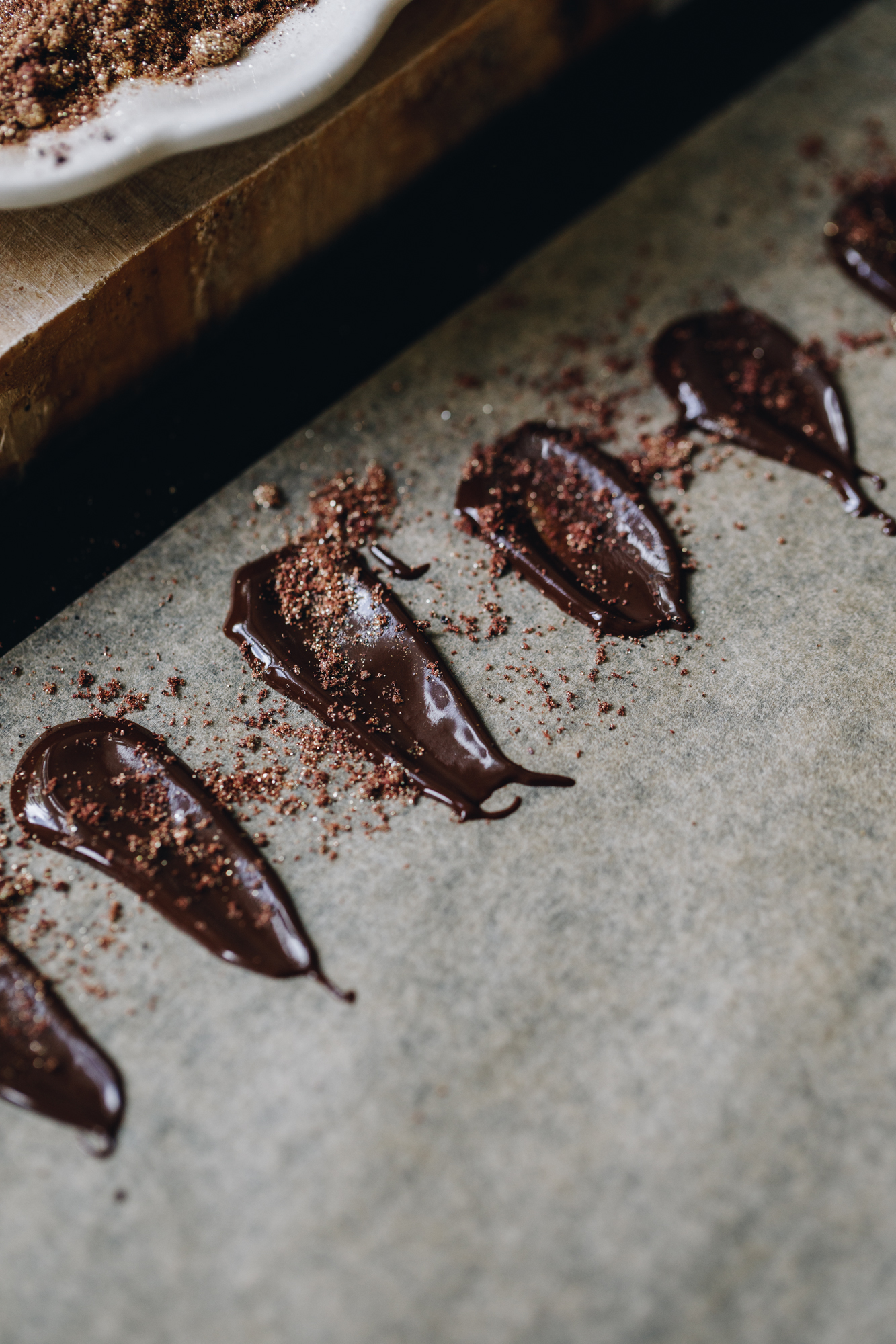 A black tray is lined with brown baking paper. On the paper, is melted chocolate that have been made in to a petal shape. They are sprinkled with fruit powder. 