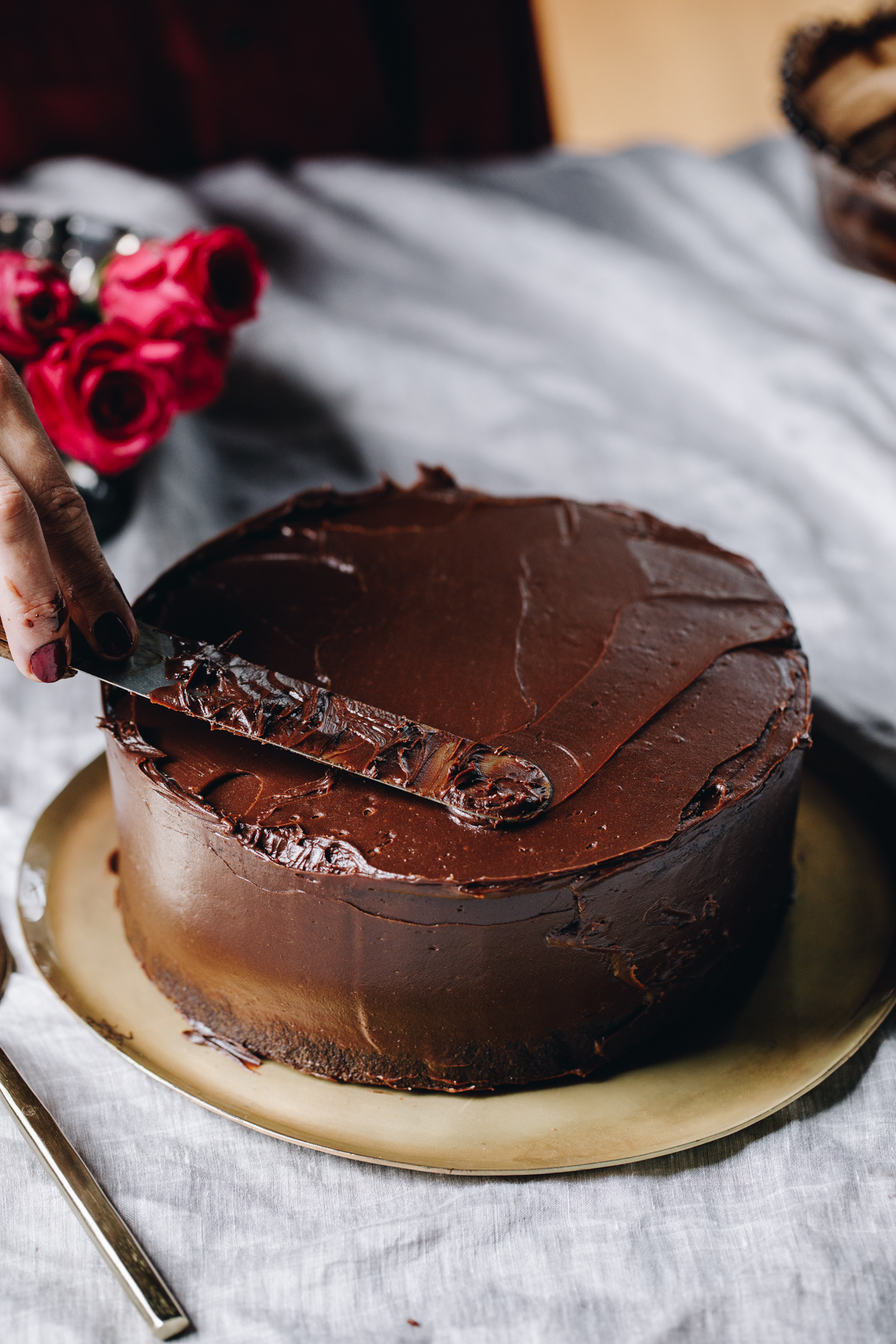 A chocolate cake sits on a golden plate on top of a grey tablecloth. The cake is being smother in a glossy chocolate ganache. Pink roses are in a vase behind it. 