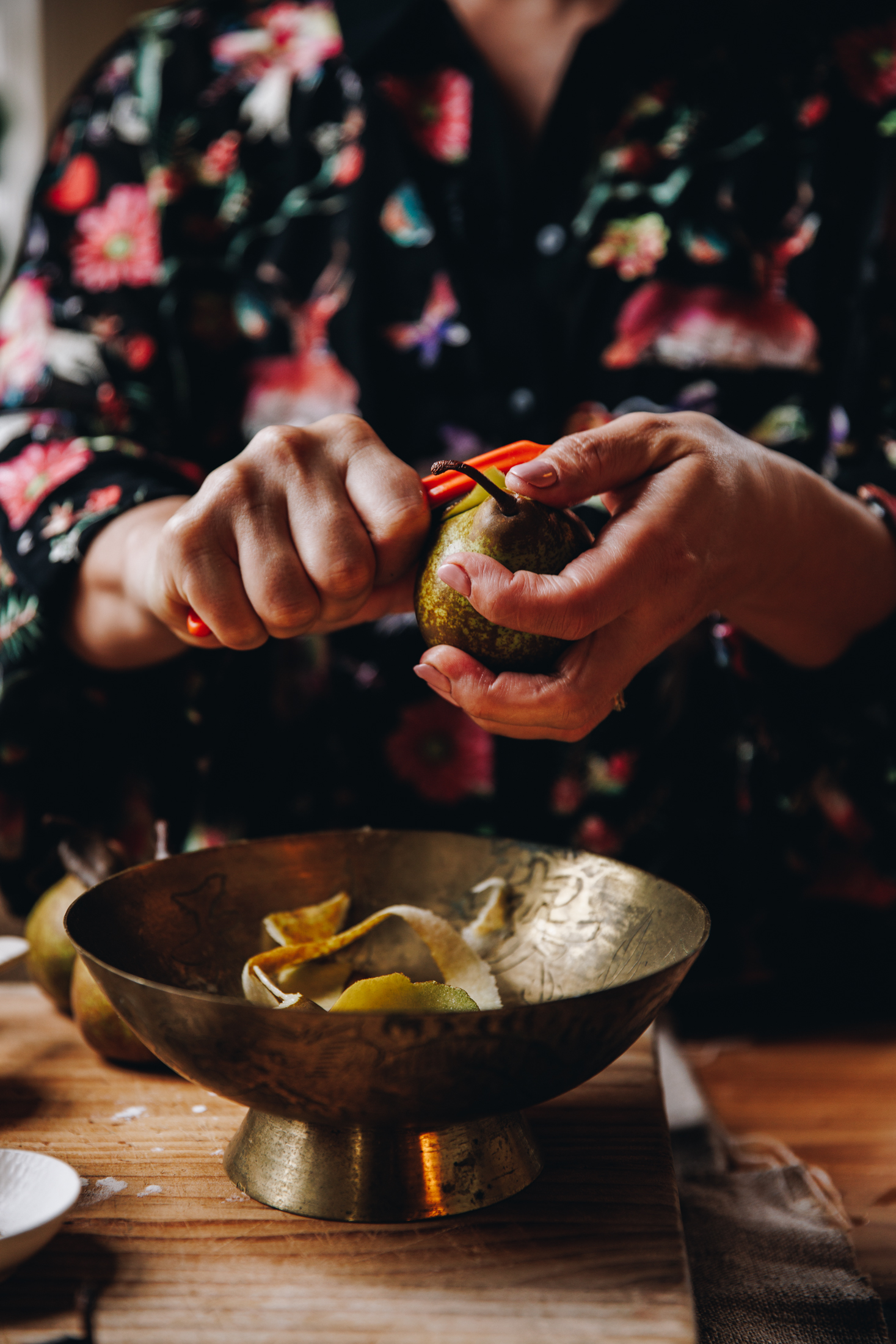 Naomi holding a pear and is peeling it with an orange potato peeler. The peels are falling in a bronze bowl that is on a wooden board that sits on a wooden table.