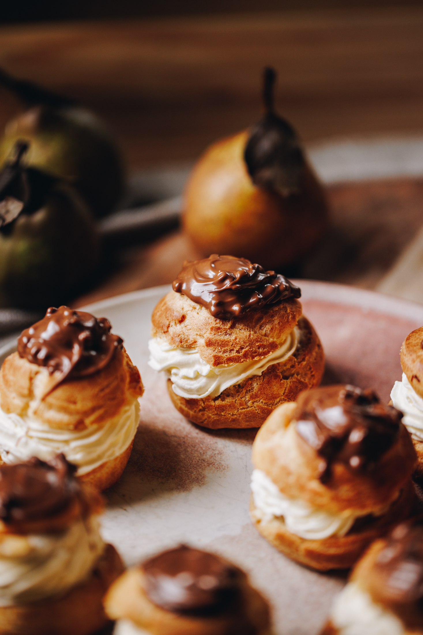 On a pink plate sits cream filled choux buns with chocolate on top. Behind the plate is three pears with leaves on them. 