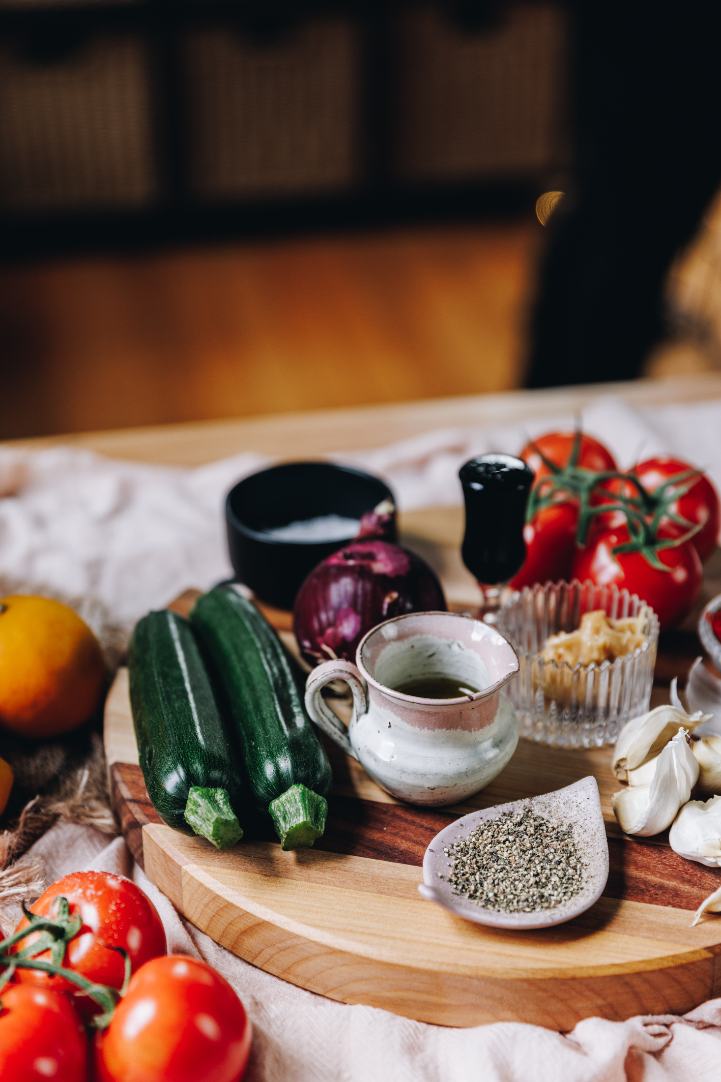 A round wooden board sits on a pink tablecloth. On it is whole courgettes and garlic cloves. Next to them is small bowls and jars filled with ingredients. There is pepper, oil and balsamic vinegar. Whole tomatoes are in the corners of the frame. 