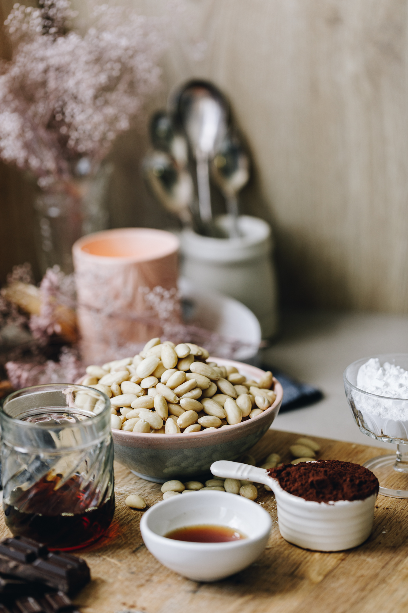 A wooden boards sits on a brown stone bench. On the board is vintage glasses, bowls and cups holding ingredients. There is vanilla, cocoa, and blanched almonds in shot. A peach candle, a white mug with spoons in it and pink dried flowers are in the background. 