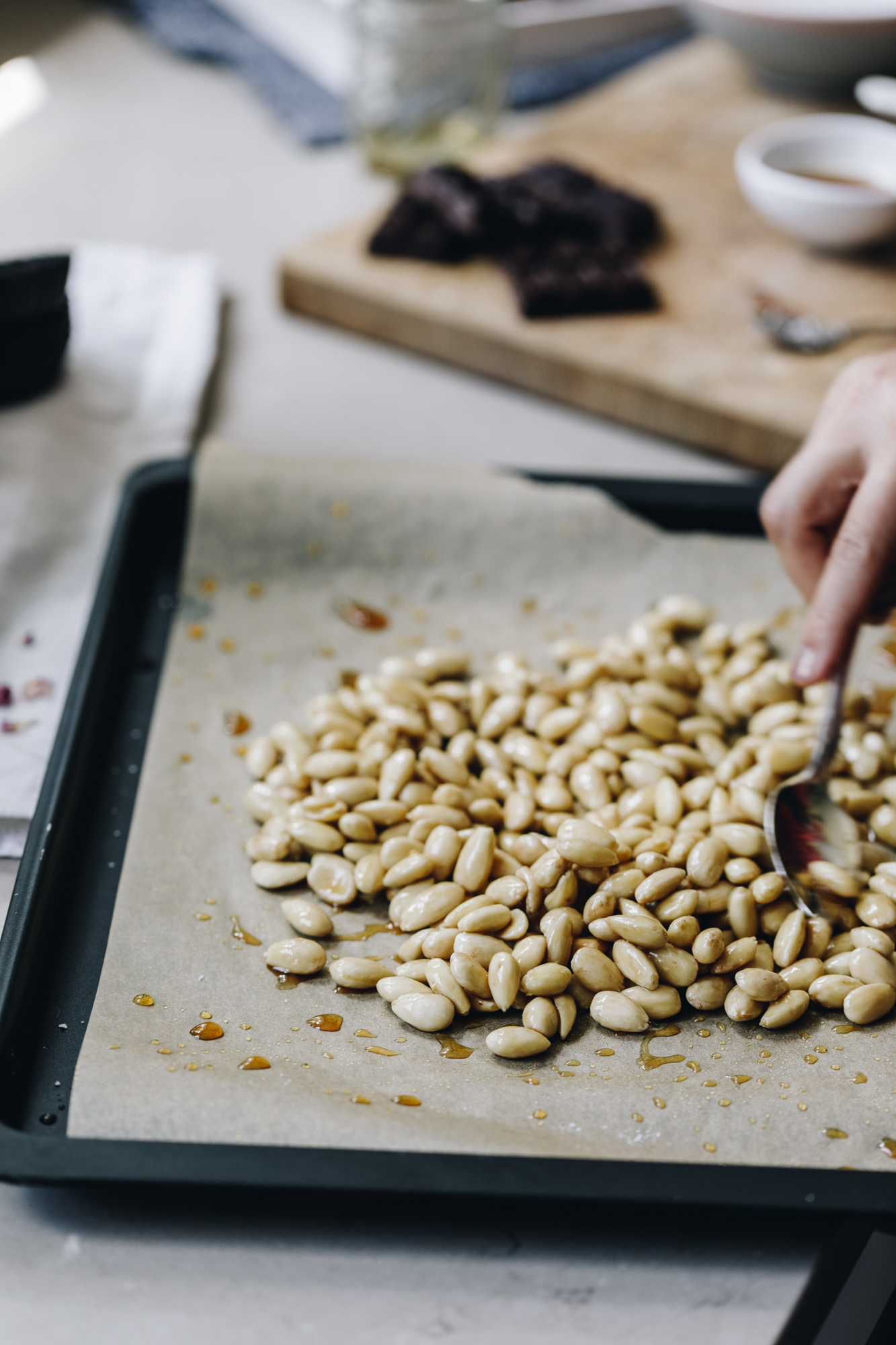 A black tray lined with brown baking paper is on a brown stone bench. On the baking paper is blanched almonds that are being stirred with a spoon. Behind the tray is a wooden board with chocolate and utensils on it. 