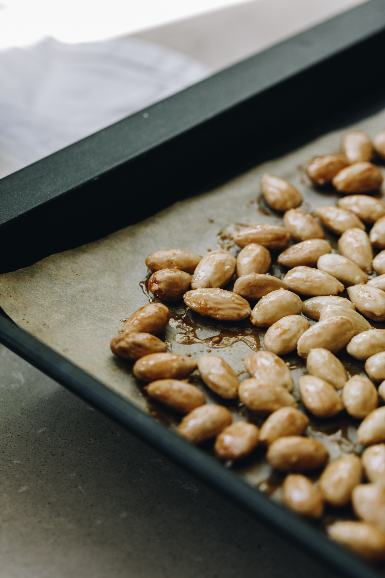 A black tray lined with brown baking paper is on a brown stone bench. On the baking paper is deeply toasted blanched almonds that are sticky with caramelised maple syrup. 