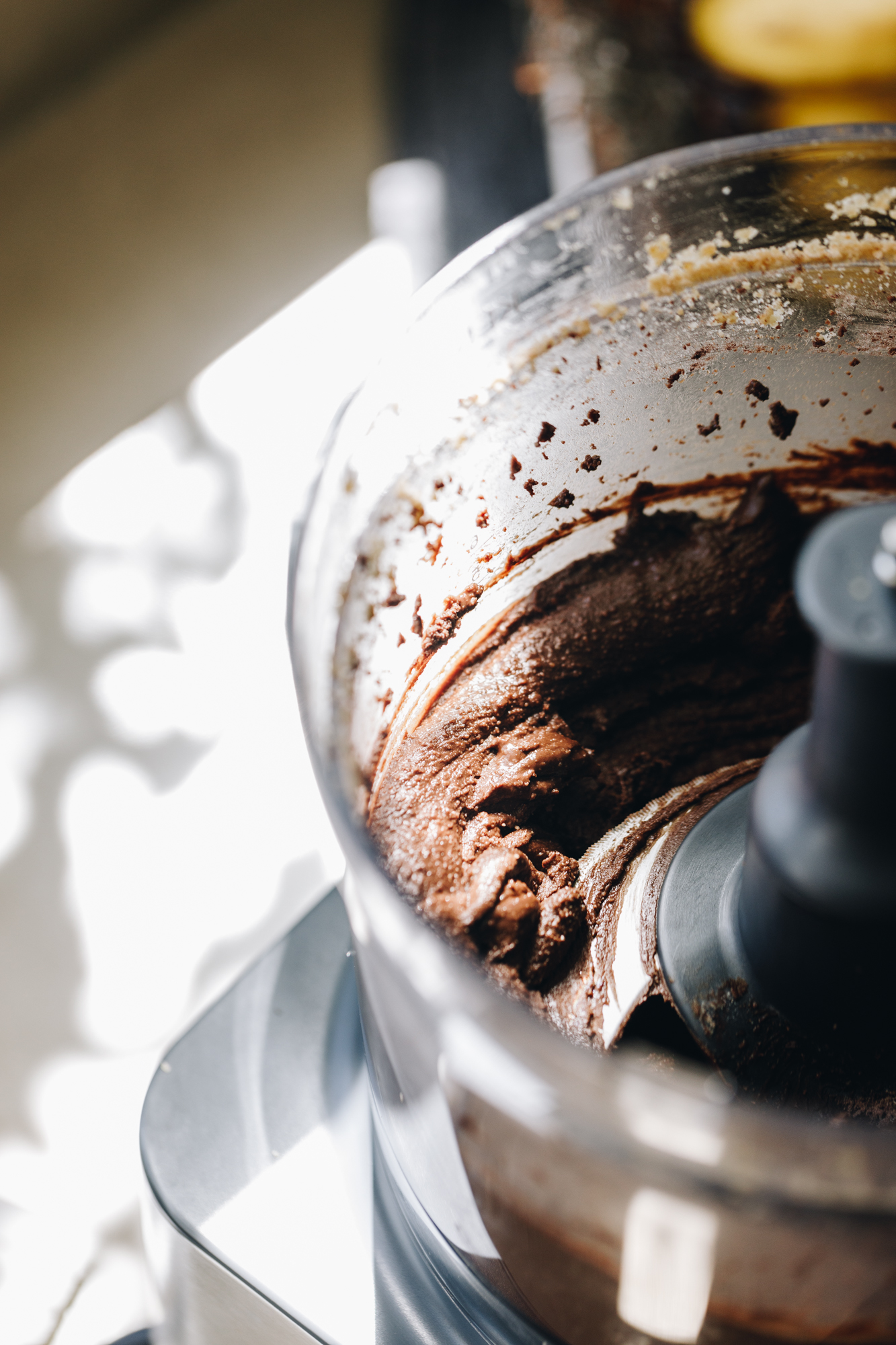 A close up shows chocolate almond butter whipped in a food processor. Speckled light is shining on the bench next to it. 
