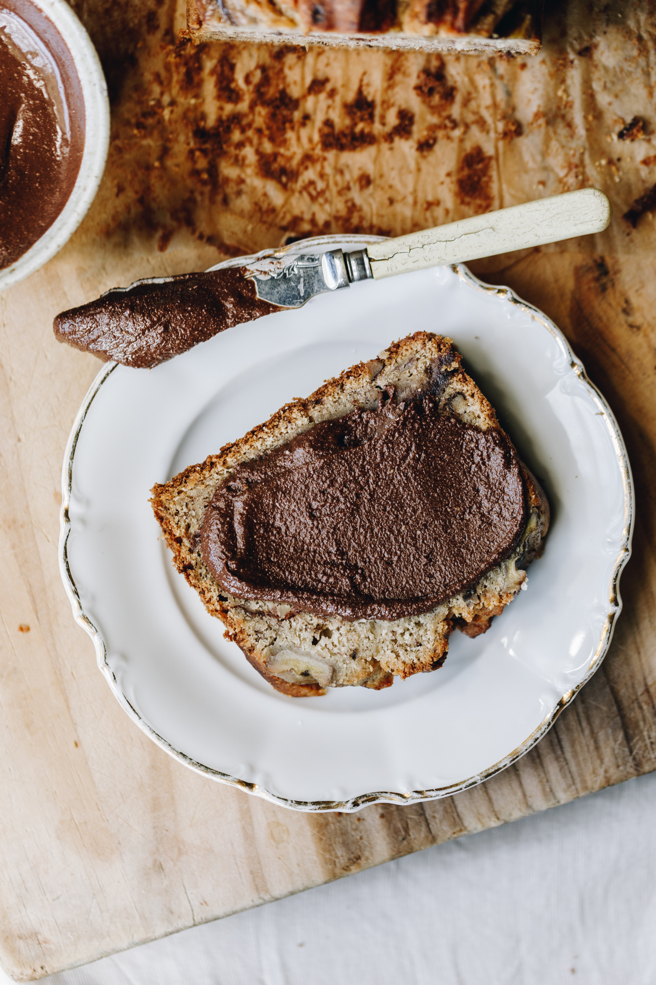 A slice of banana loaf is on a white vintage plate. On top of it is a thick layer of chocolate almond butter with a knife next to it with more chocolate almond butter on it. The plate is on a wooden board with brown baking paper and a glimpse of a banana loaf and more almond butter. 