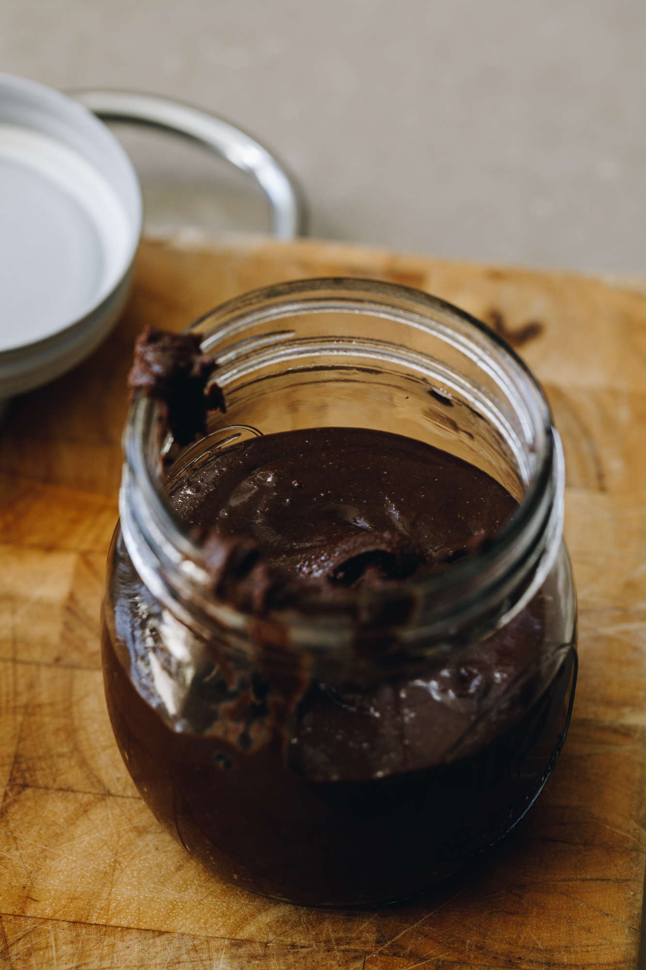 A small wooden board sits on a brown stone bench. On the board is a small glass jar with chocolate almond butter in it. The white lid sits next to it. 