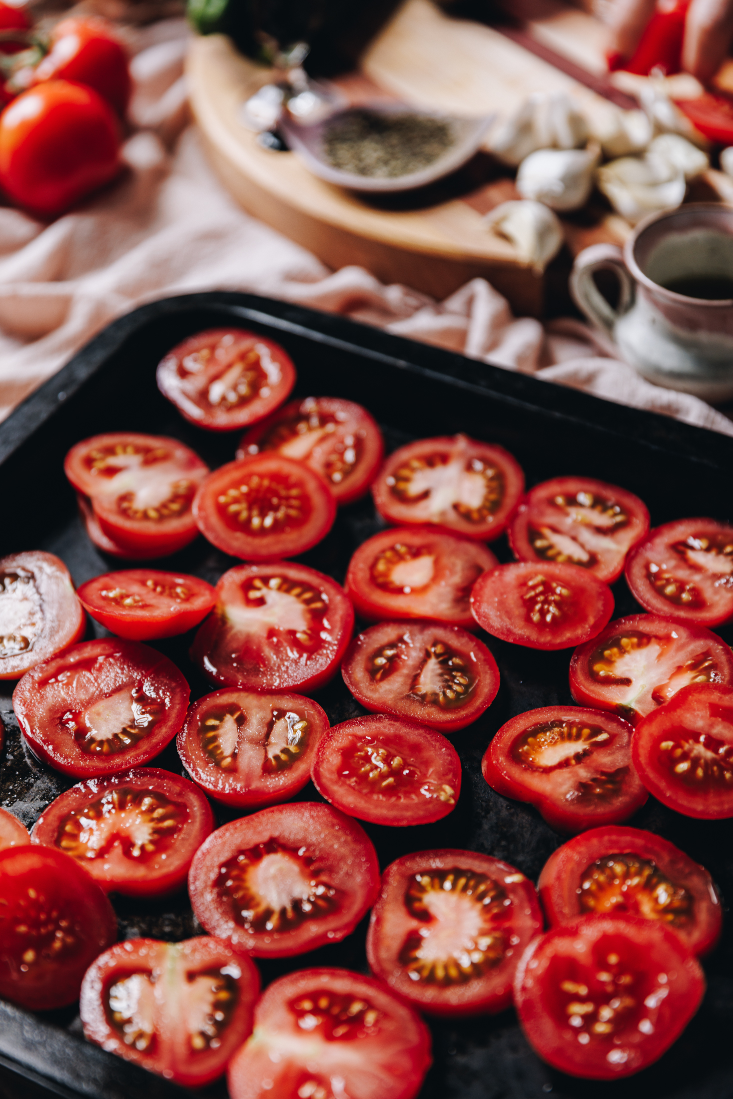 A black oven tray is sitting on a pink tablecloth. In the tray is slices of fresh tomatoes. Behind the tray is a round board with more ingredients of garlic cloves, pepper, a small jug and more tomatoes. 