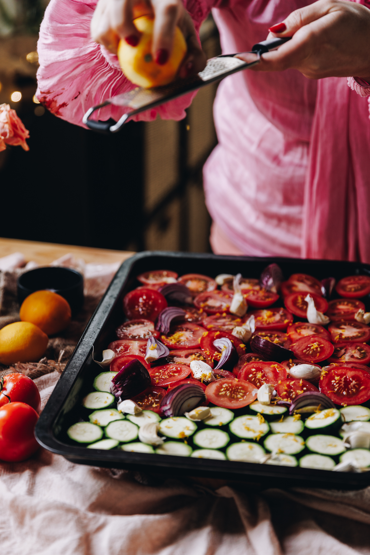 A black oven tray is sitting on a pink tablecloth. In the tray is slices of fresh tomatoes, sliced courgettes, sliced red onion and garlic cloves. Naomi is using a silver grater to grate lemon zest on top of the vegetables. More tomatoes and lemon are next to the tray.