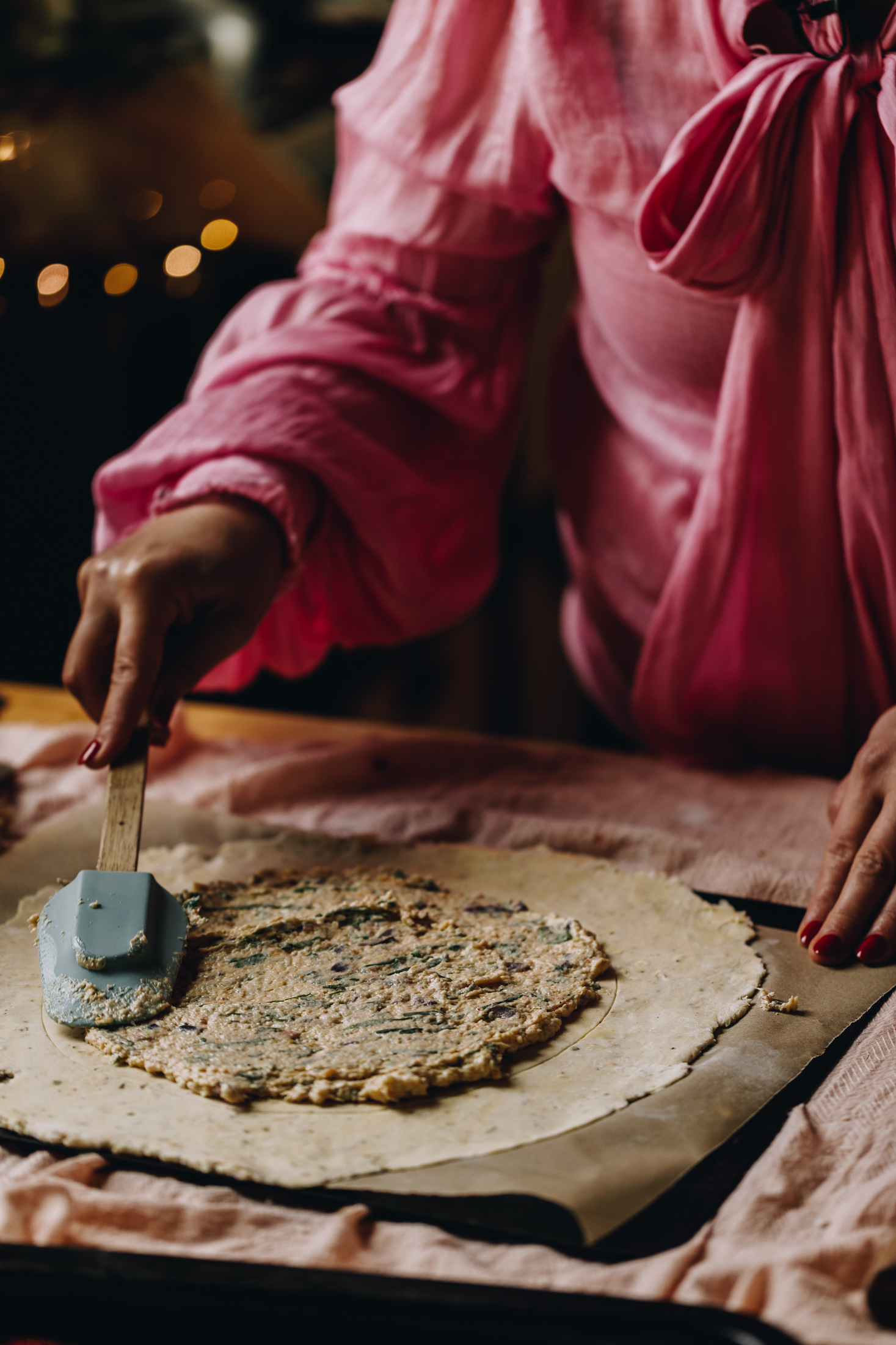A light pink tablecloth lines a table. On the cloth is a black tray lined with brown baking paper. On the baking paper is a pastry that has been rolled in to a circle. Naomi is spreading a creamy garlic cream on to the centre with a spatula. 
