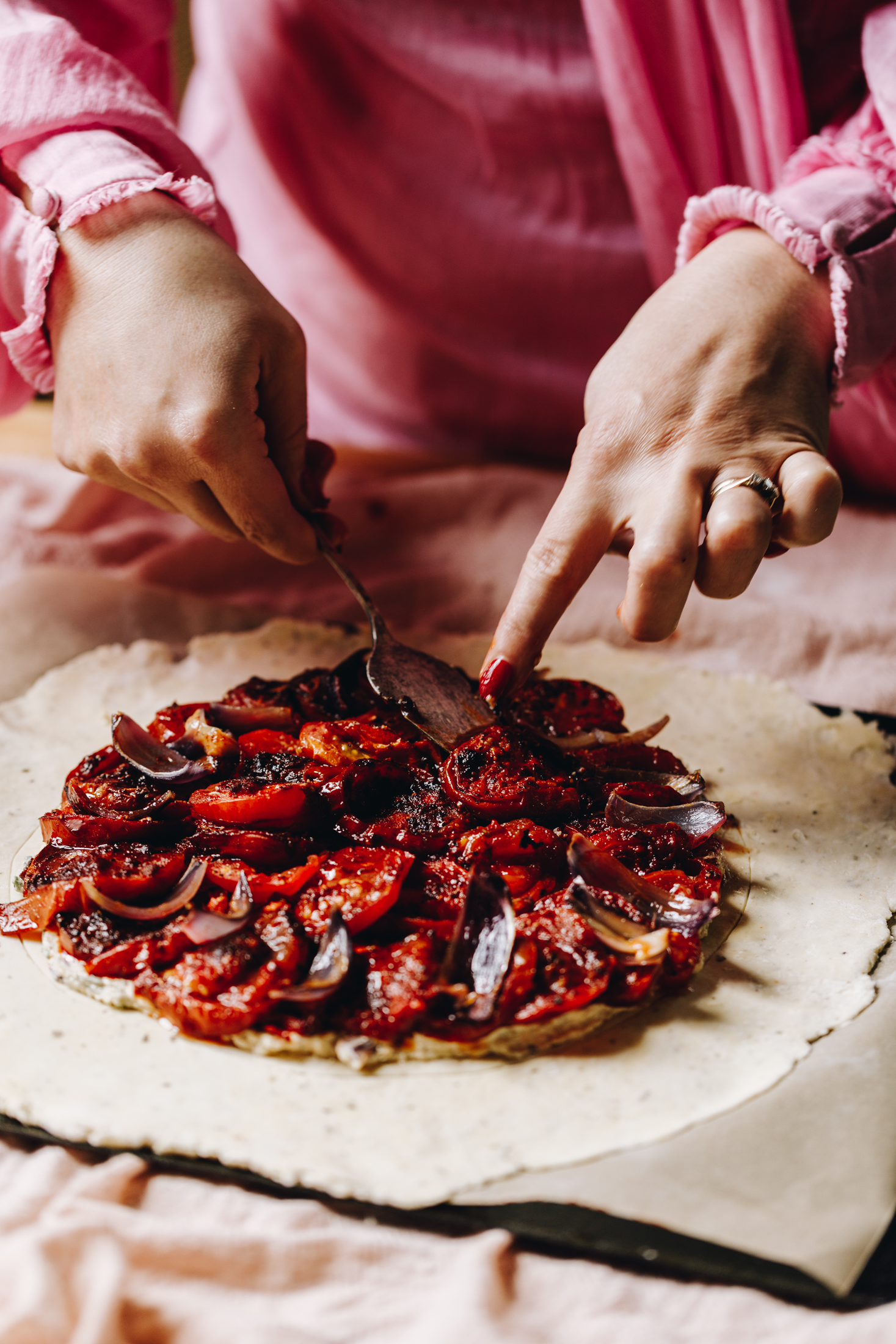 A light pink tablecloth lines a table. On the cloth is a black tray lined with brown baking paper. On the baking paper is a pastry that has been rolled in to a circle. Naomi is placing a roasted tomato on top in the centre which is part of pile of roasted tomatoes, courgettes and red onion. 
