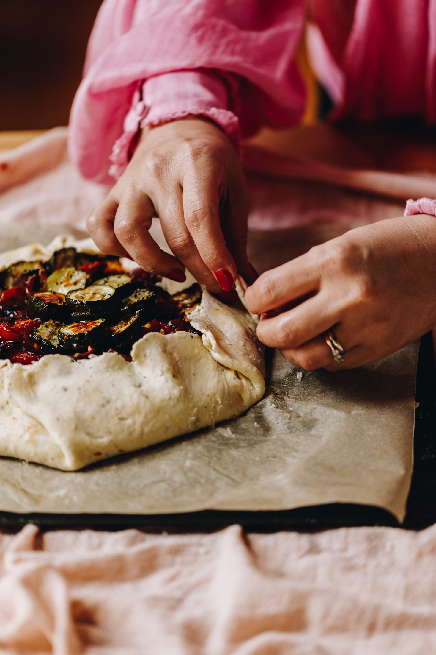 A light pink tablecloth lines a table. On the cloth is a black tray lined with brown baking paper. On the baking paper is a pastry that has been filled with roasted tomatoes and courgettes. Naomi is holding the pastry and folding the edges in to encase the filling. 