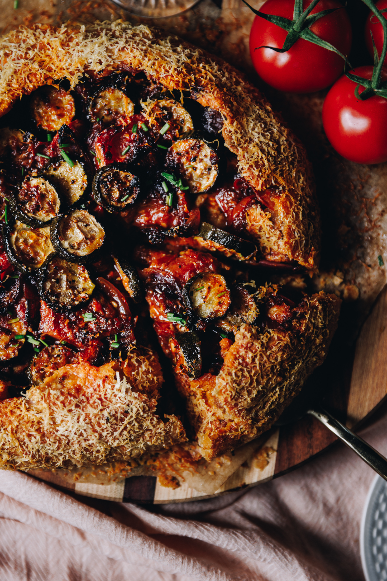 A flat lay shot shows a tomato galette that is wrapped in herbed shortcrust savoury pastry. Fresh tomatoes can be seen in the top left corner. The galette is hole but one piece has been cut and a cake server is lifting it from the board. 