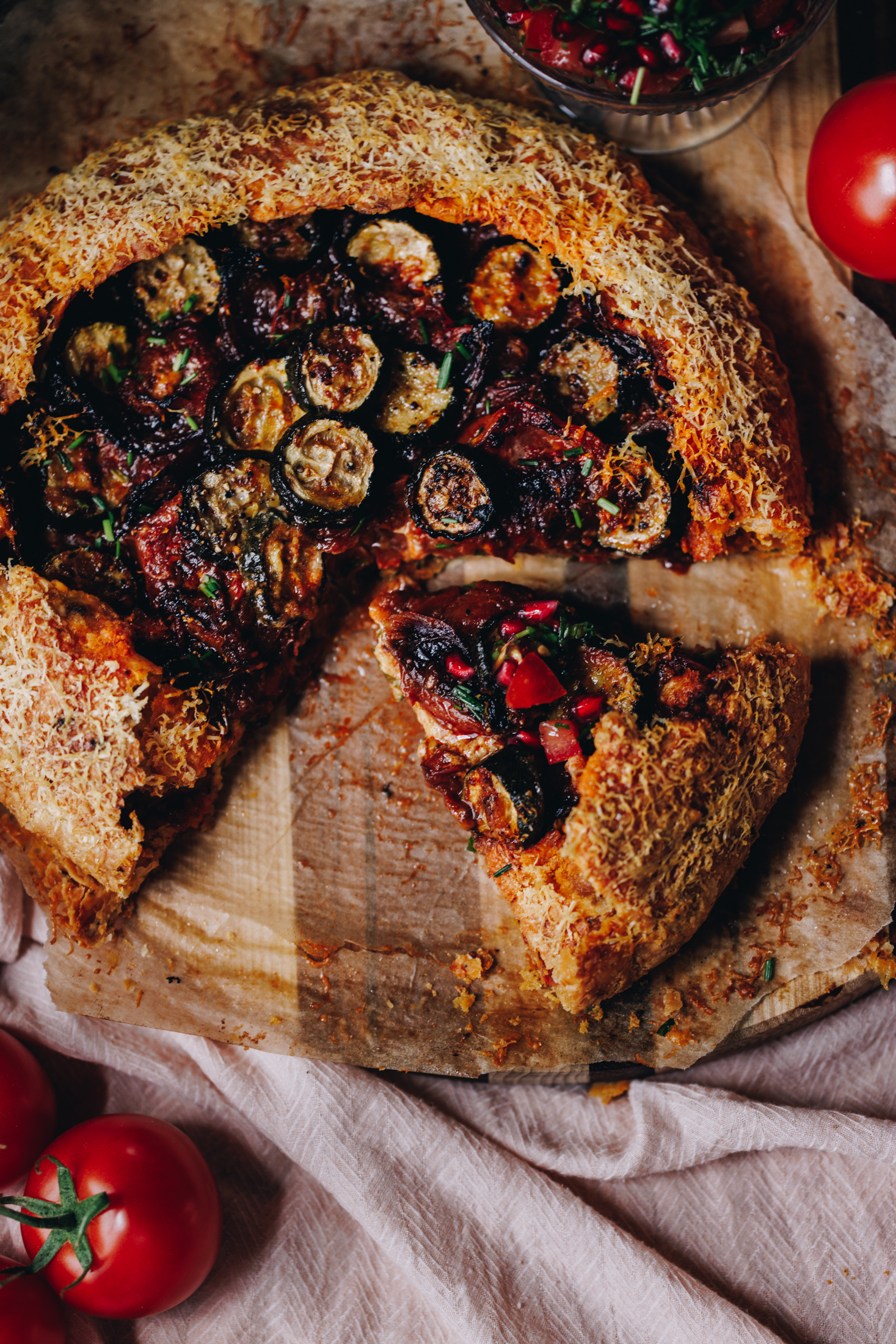 A flat lay shot shows a roasted tomato and courgette galette that is wrapped in herbed shortcrust savoury pastry. Fresh tomatoes can be seen in the top left corner. The galette is hole but there is a piece removed and one piece is cut. 