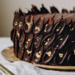 A chocolate cake is decorated with is decorated with dark chocolate petal decorations, each one has gold leaf on them.