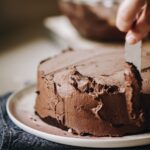 A small chocolate cake is being spread with vegan chocolate buttercream with a knife. The cake is on a pink plate.