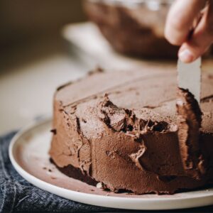 A small chocolate cake is being spread with vegan chocolate buttercream with a knife. The cake is on a pink plate.