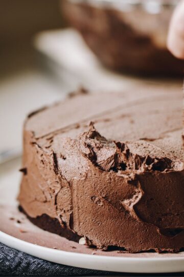 A small chocolate cake is being spread with vegan chocolate buttercream with a knife. The cake is on a pink plate.